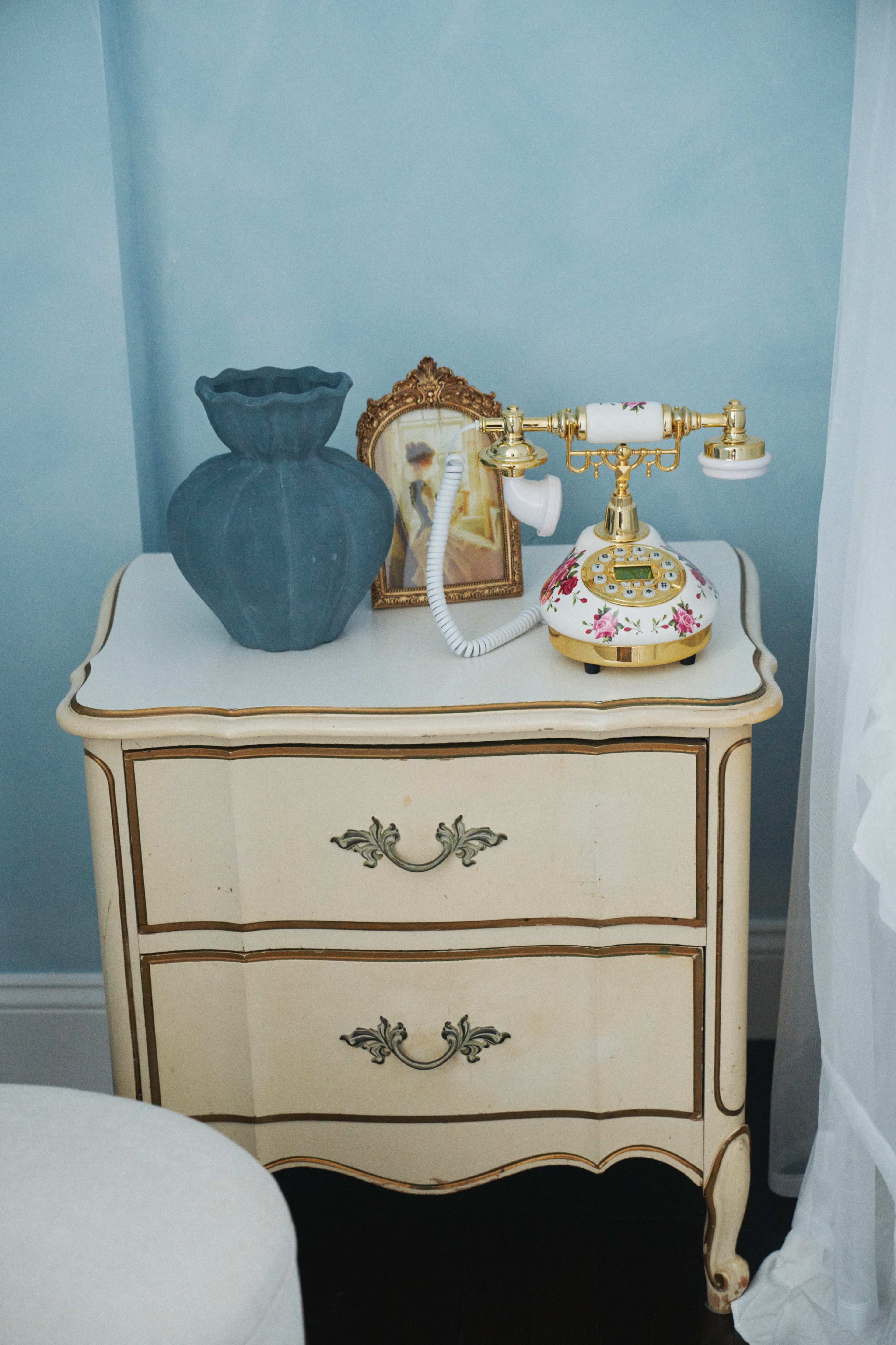 A vintage nightstand holds a decorative vase, a framed picture, and an ornate telephone against a light blue wall.