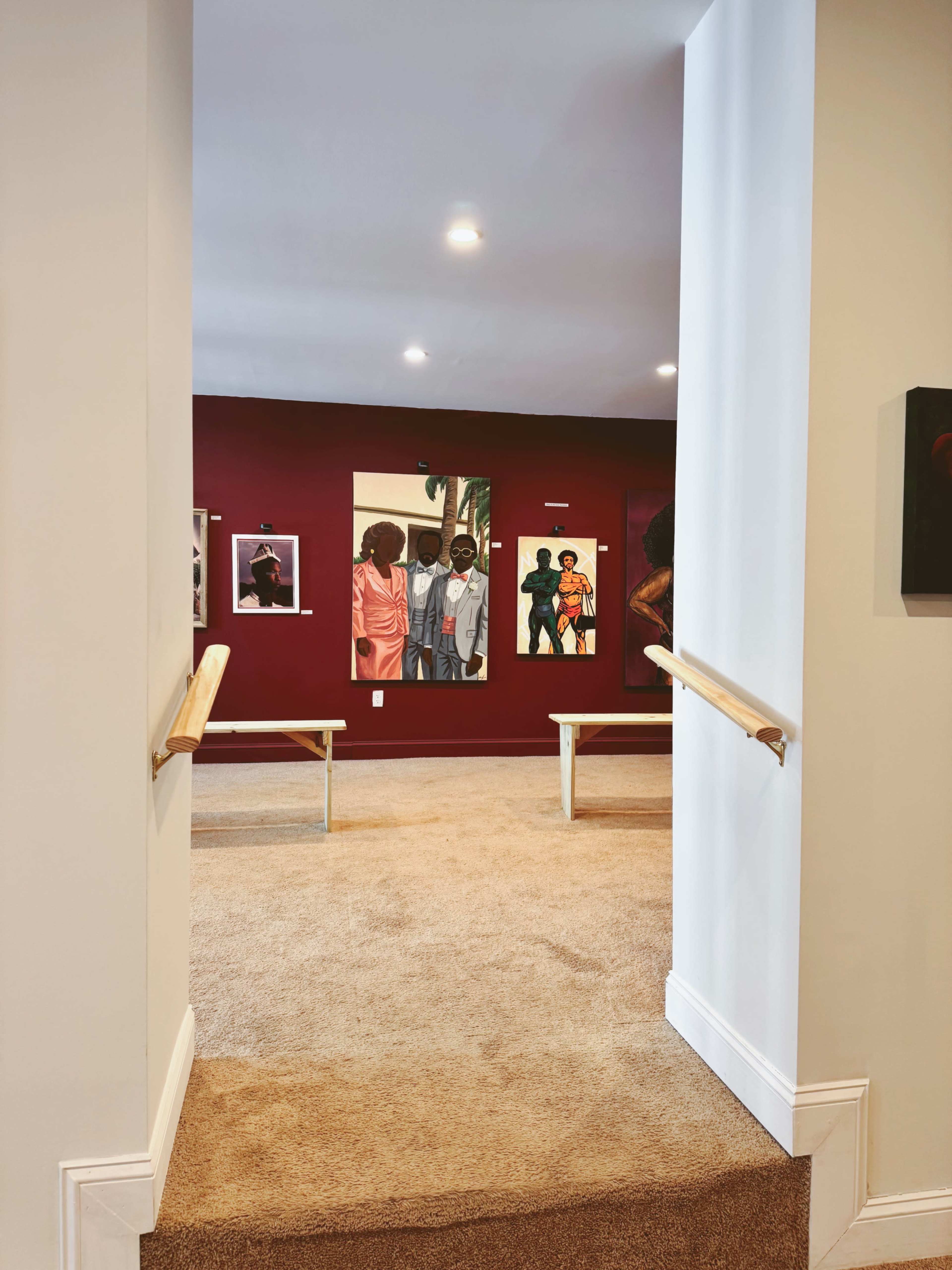The image shows a view from an entryway into a gallery space with burgundy walls displaying framed artworks and two benches in the foreground.