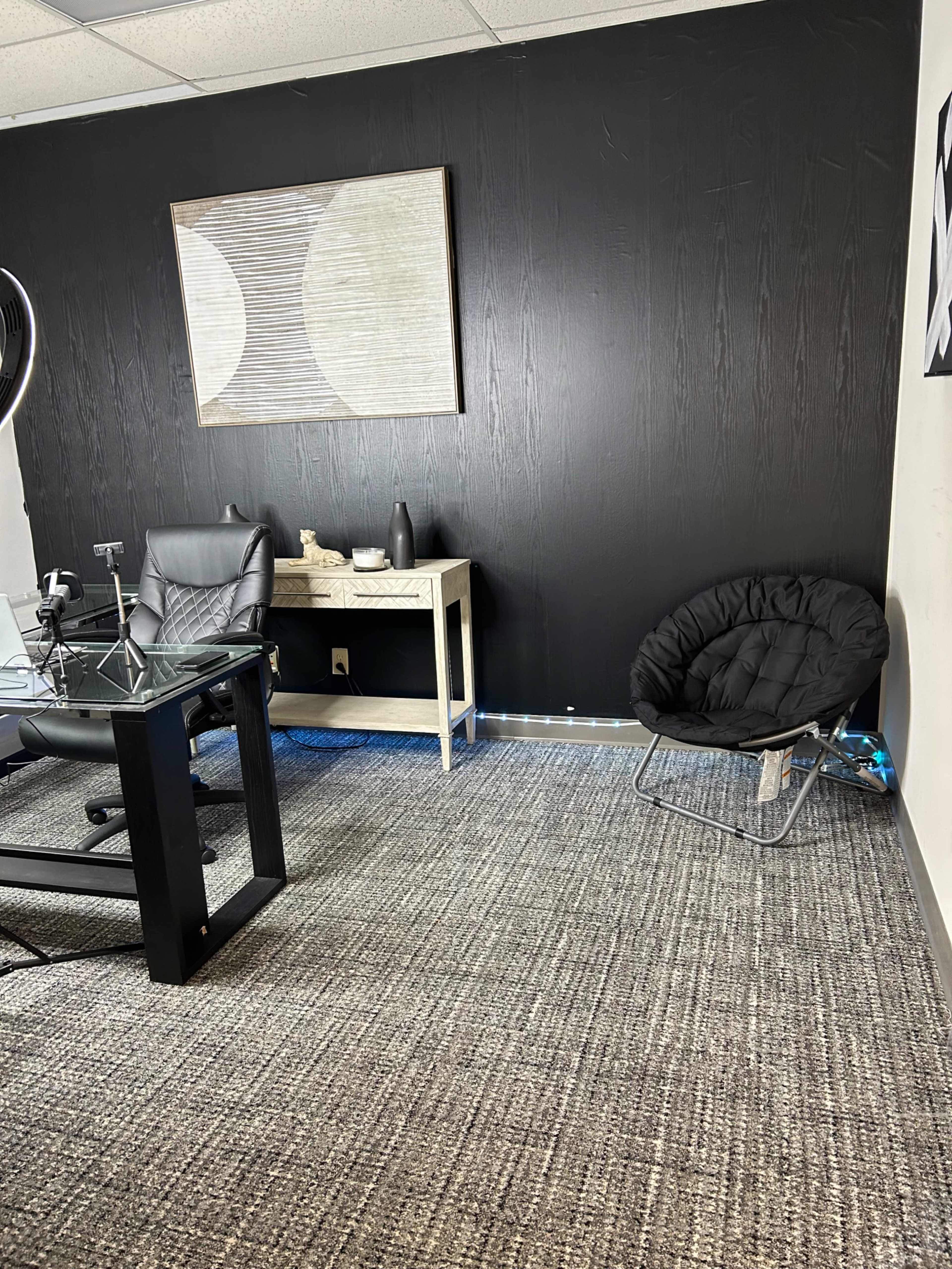 A modern office space featuring a black wall, a glass desk with a black ergonomic chair, and a round black chair in the corner.
