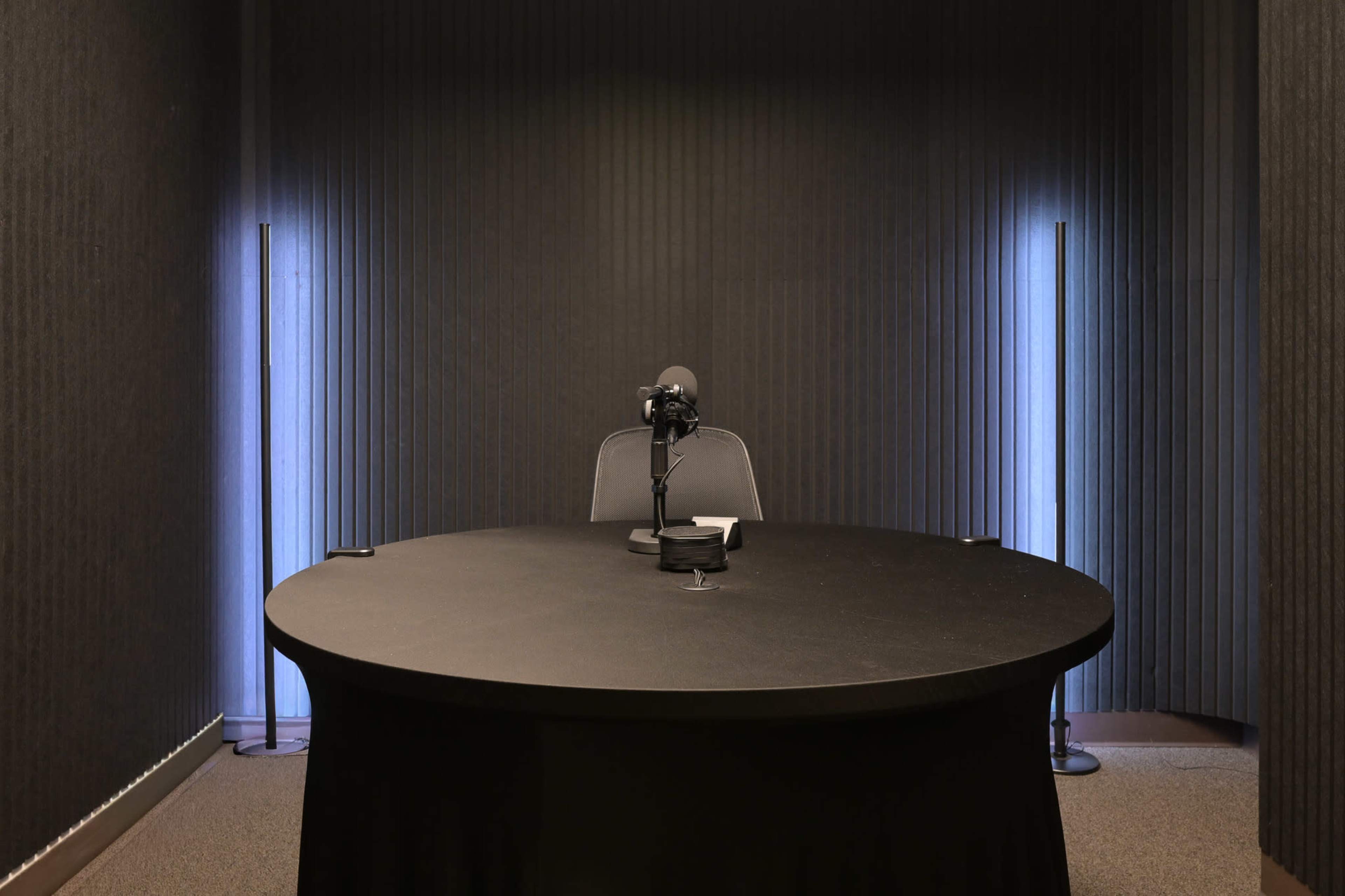 A small, dimly lit recording studio features a round table with a microphone and a chair, flanked by two tall, illuminated stands.
