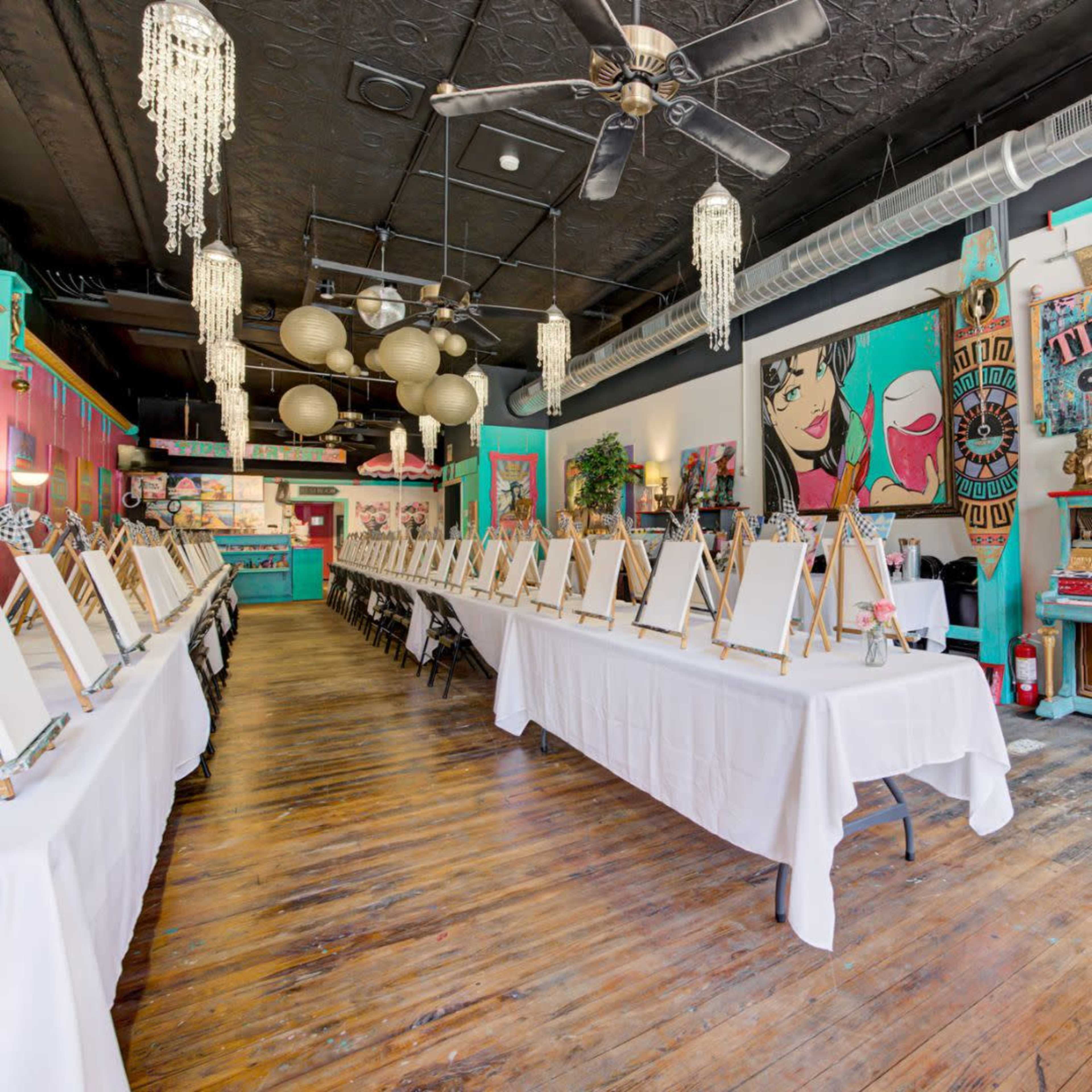 The image shows an art studio with rows of blank easels set up on tables covered with white cloth, surrounded by colorful decor and vibrant artwork.