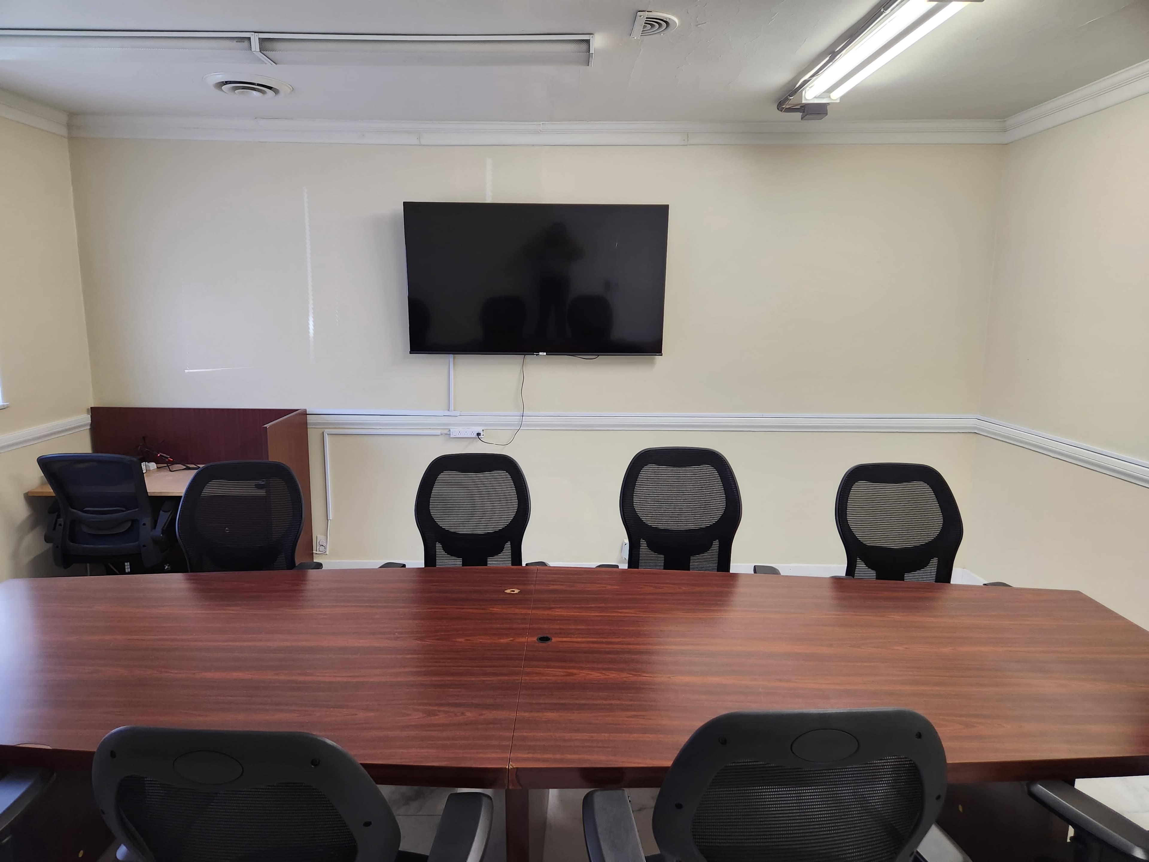 A conference room features a large wooden table surrounded by six swivel chairs and a wall-mounted television.