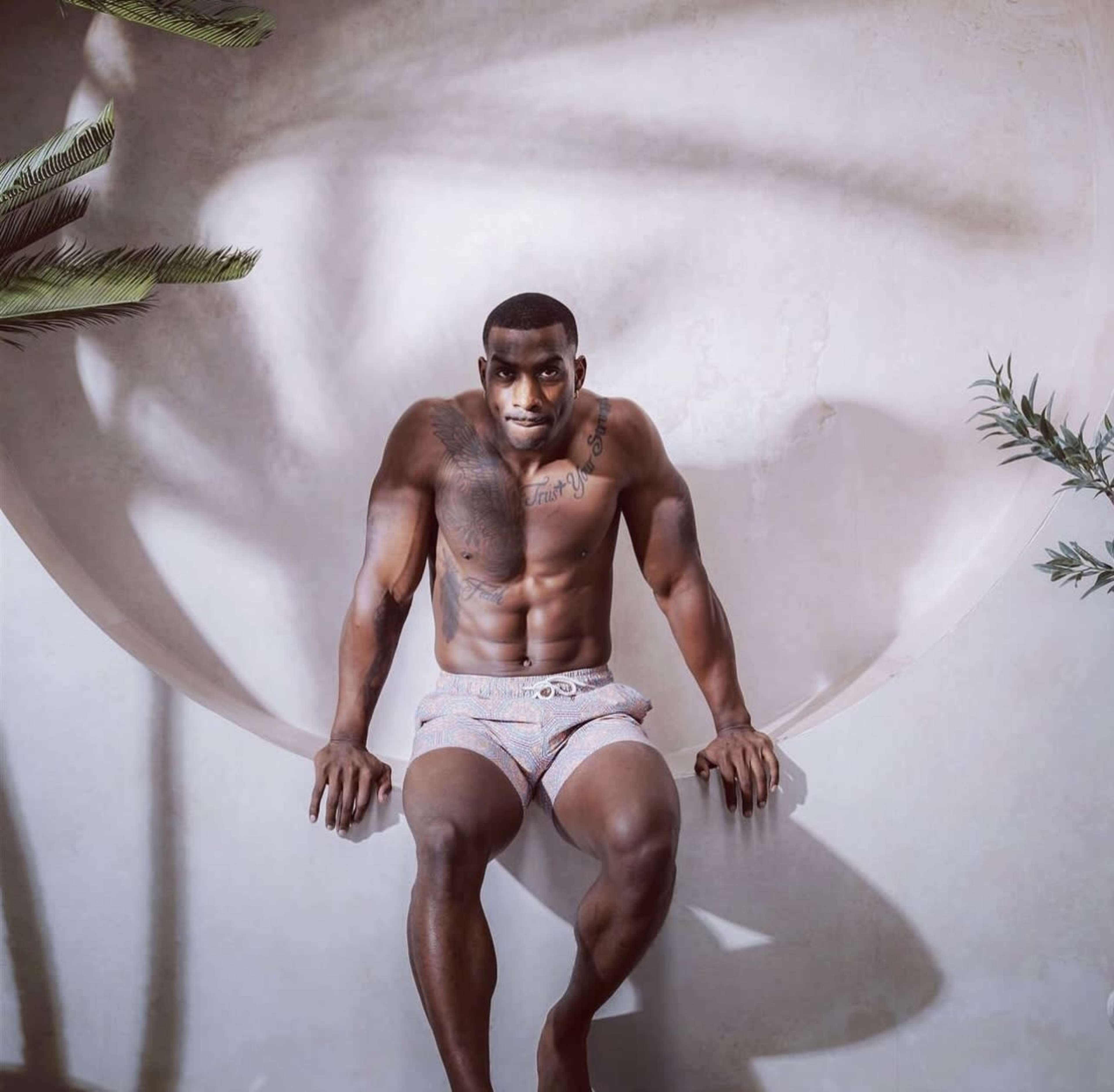A muscular man sits on a circular structure against a textured wall, surrounded by plants.