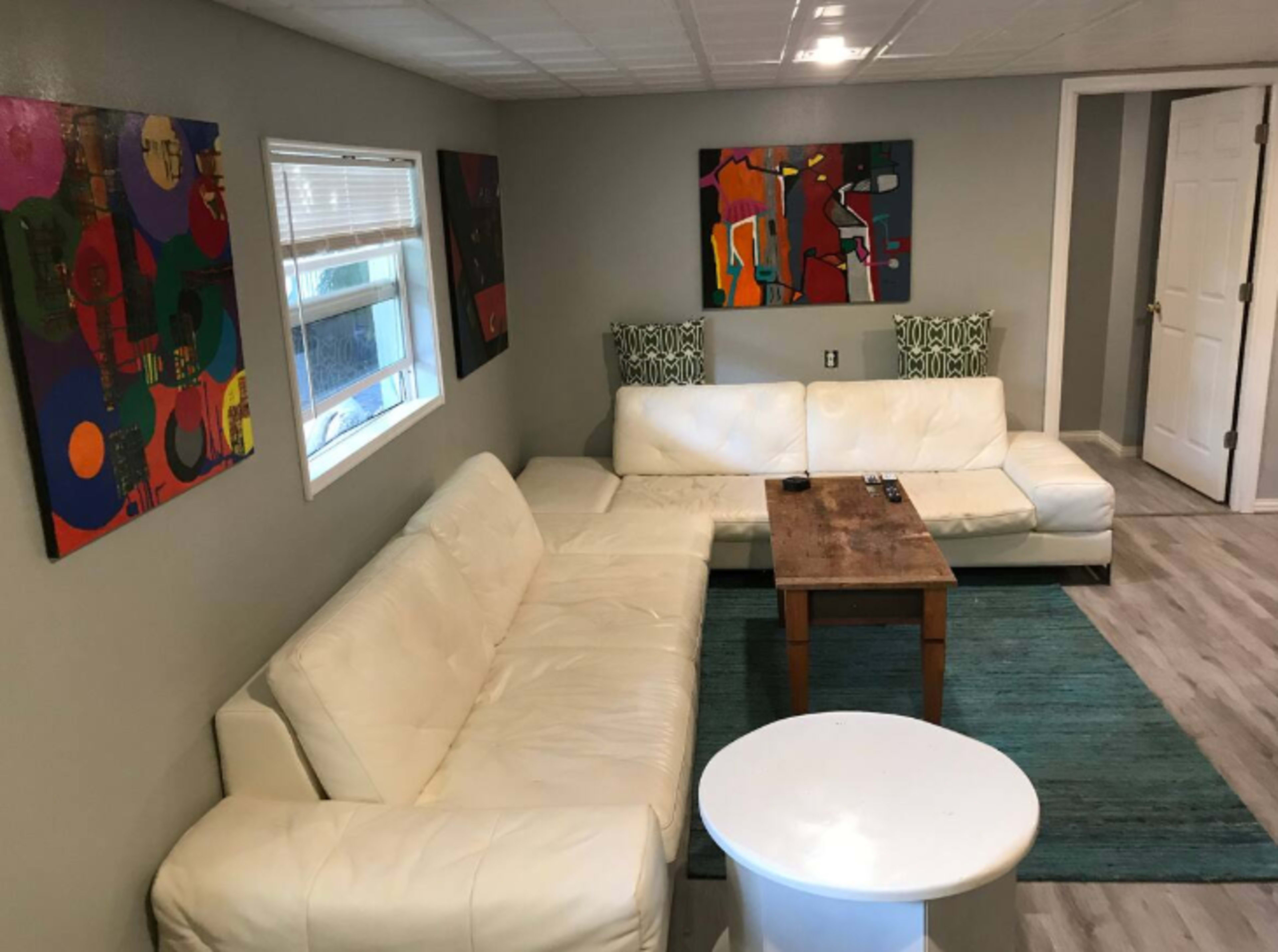 The image shows a living room with a white sectional sofa, a wooden coffee table, a round white side table, and colorful abstract artwork on the walls.
