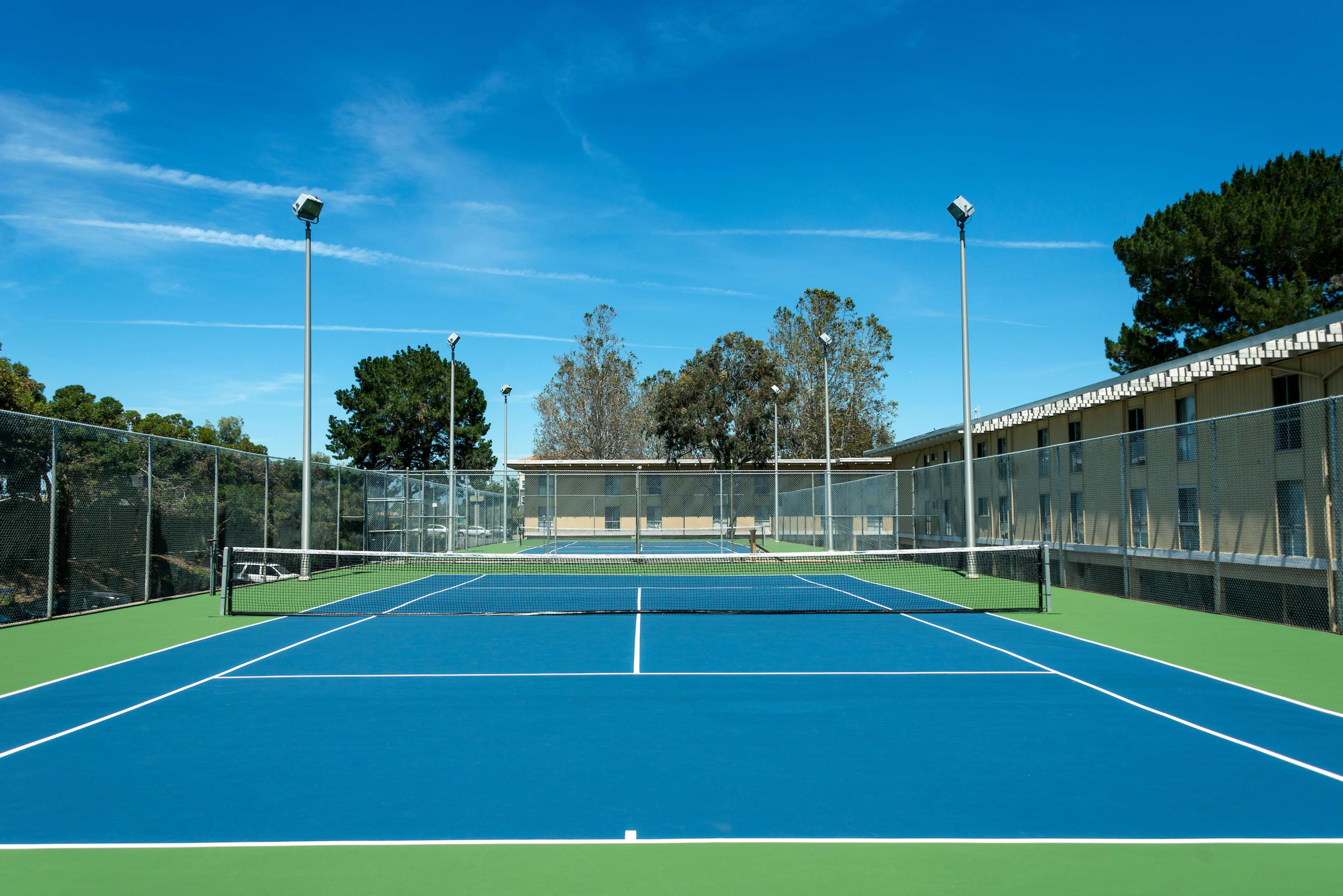 Two empty tennis courts with bright blue surfaces, surrounded by a fence and trees under a clear blue sky.