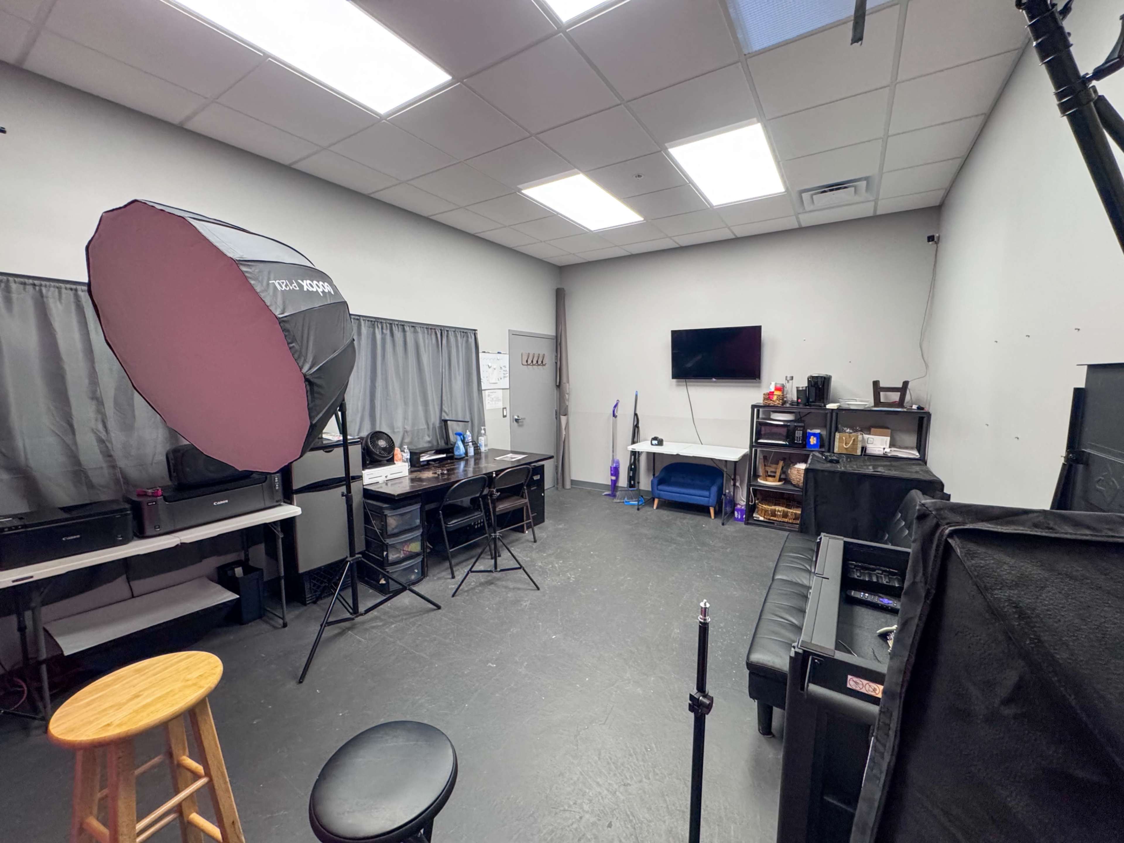 The image shows a sparsely furnished studio room containing a backdrop, lighting equipment, a television, and several workstations.