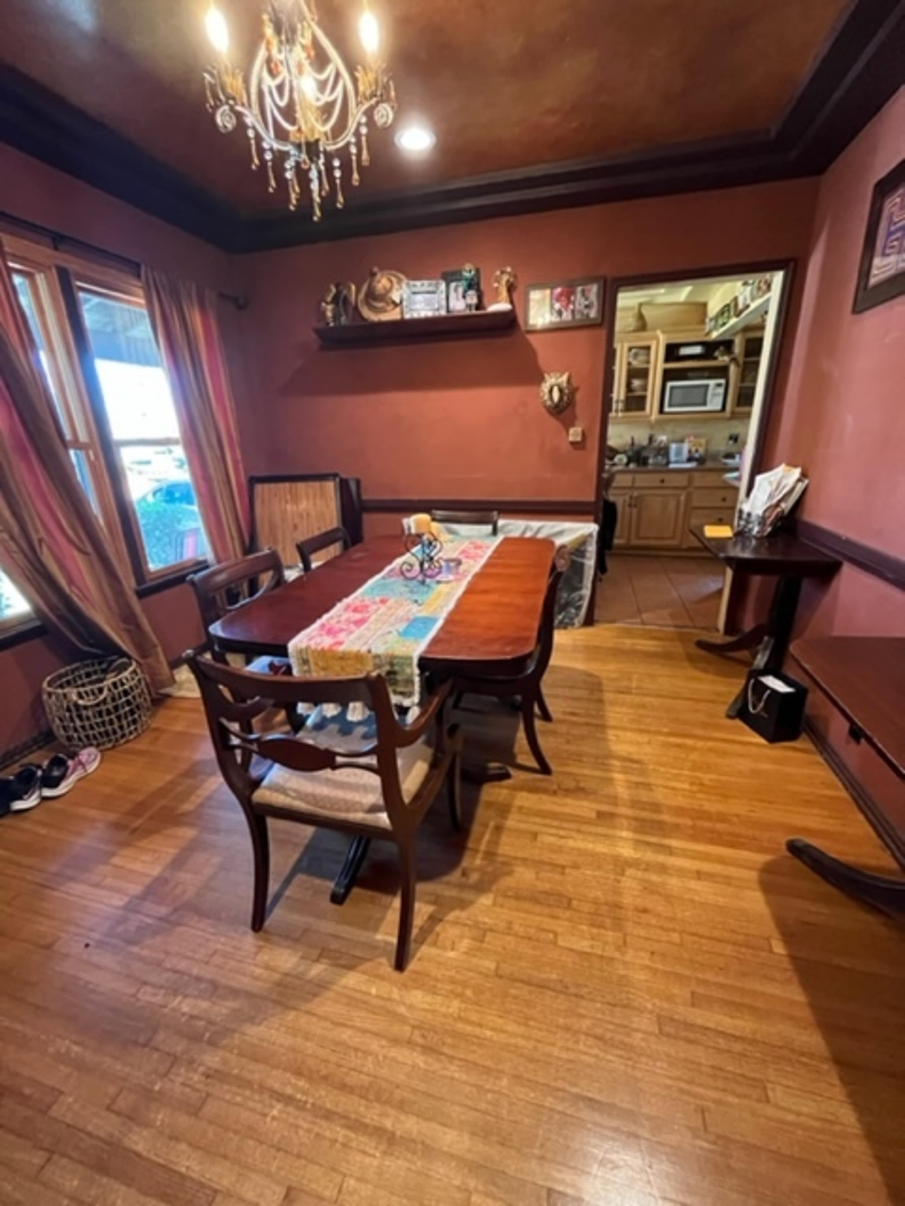 The image shows a dining area with a wooden table surrounded by chairs, featuring a colorful tablecloth and framed decorations on the walls.