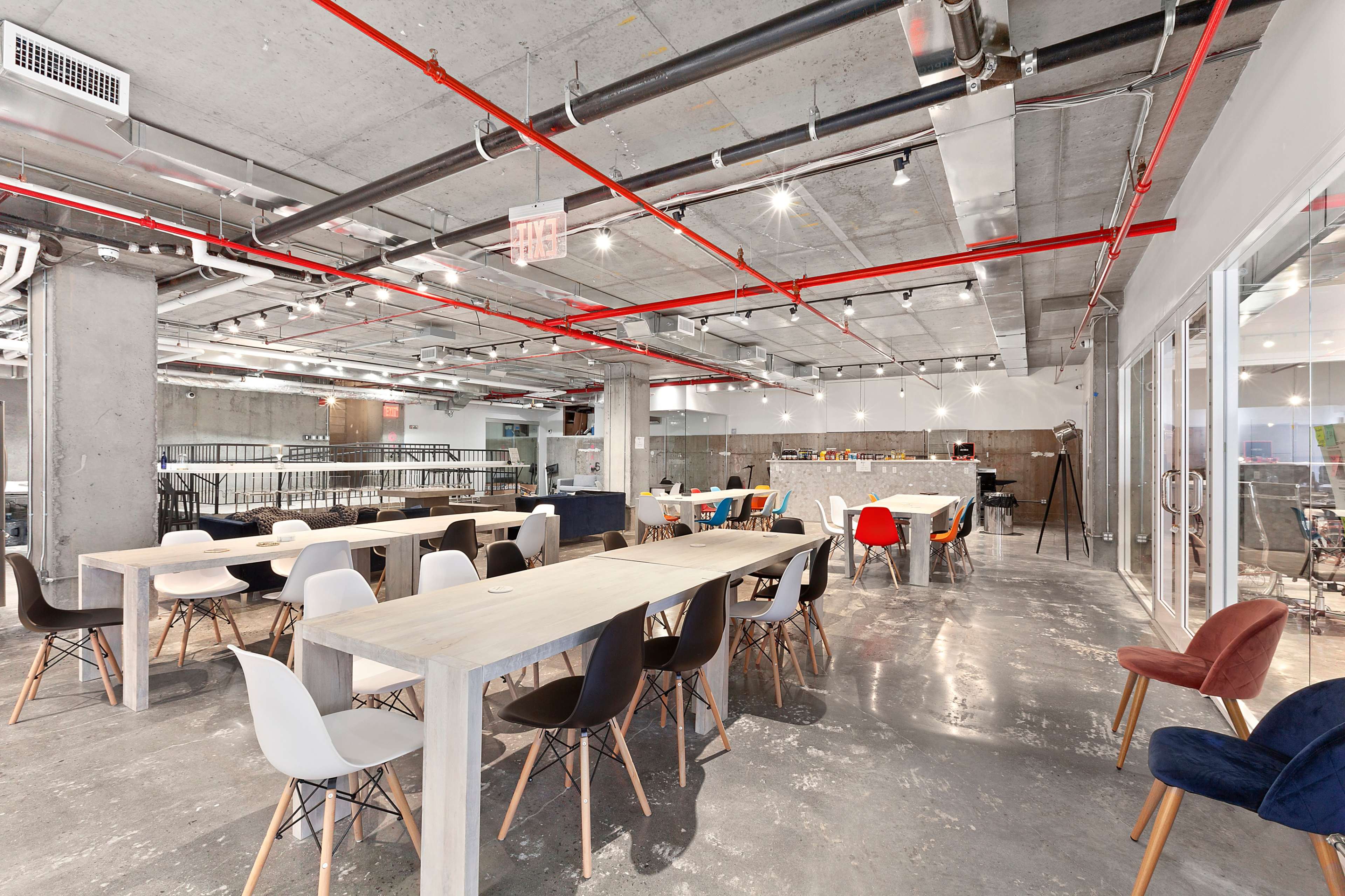 The image shows a modern, industrial-style open workspace with long tables, colorful chairs, and exposed pipes in the ceiling.