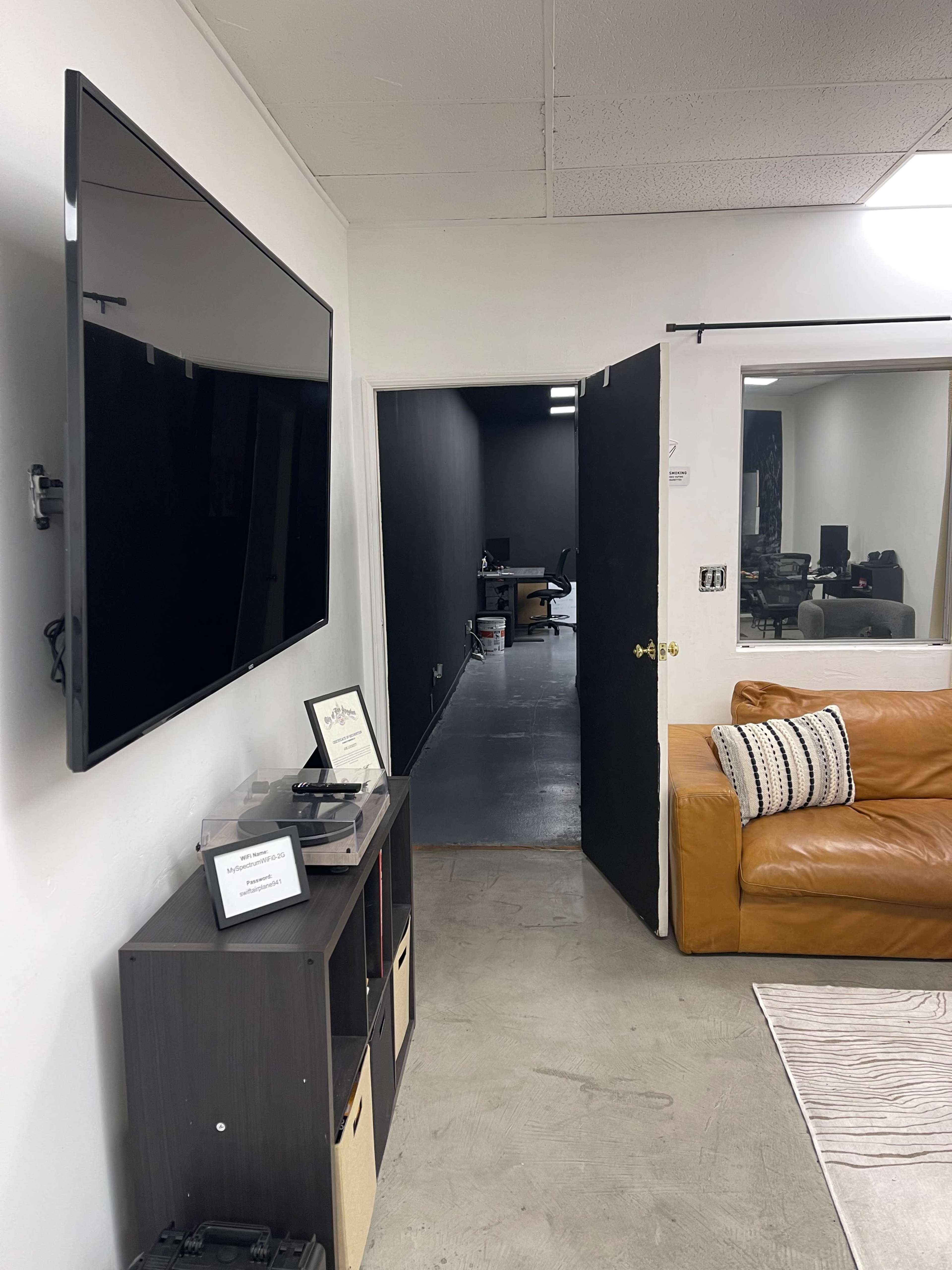 A modern interior with a wall-mounted television, a brown sofa, and an open door leading to a dark hallway with a workspace visible at the end.