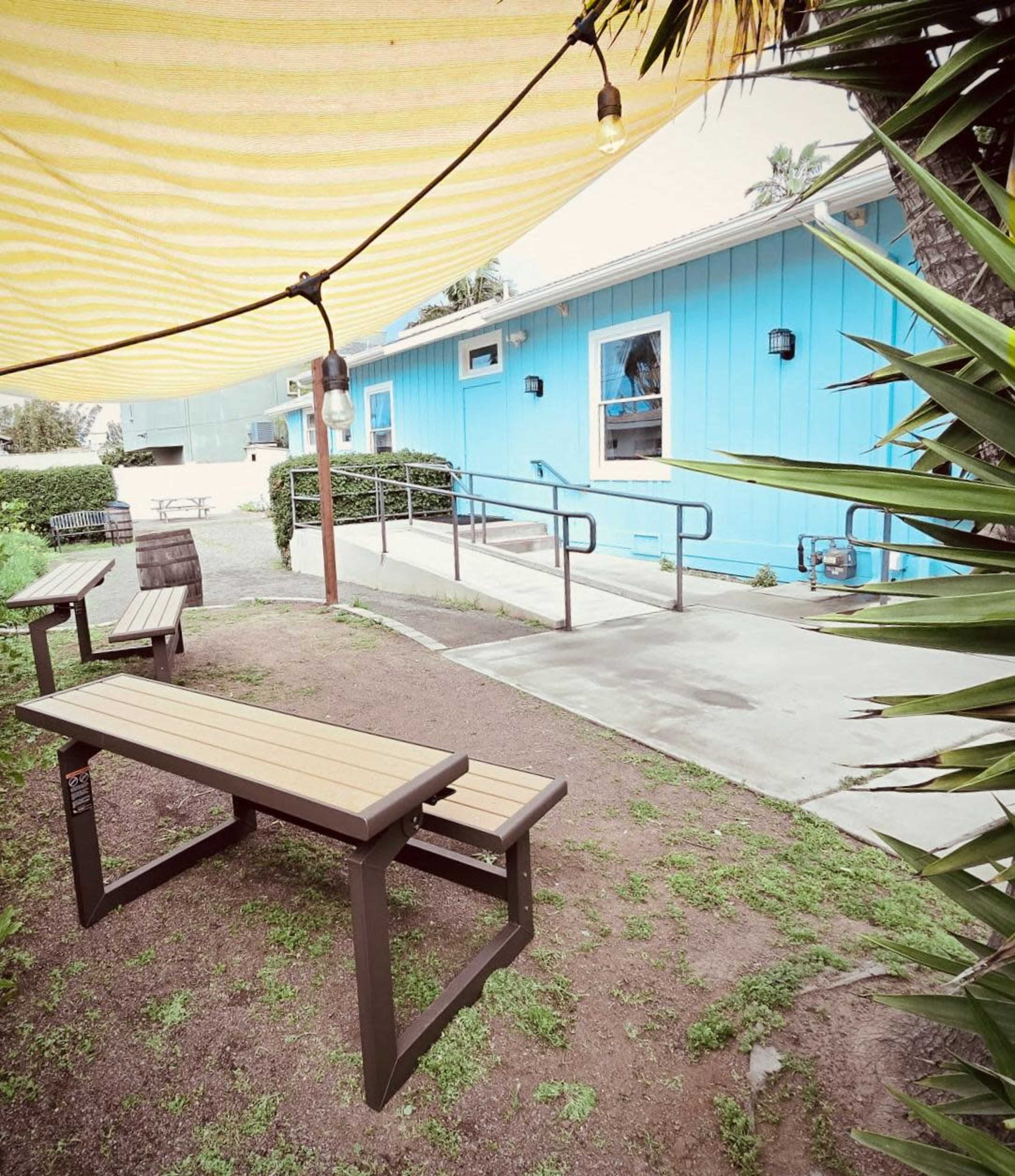 The image shows a shaded outdoor seating area with wooden benches and a blue building in the background.