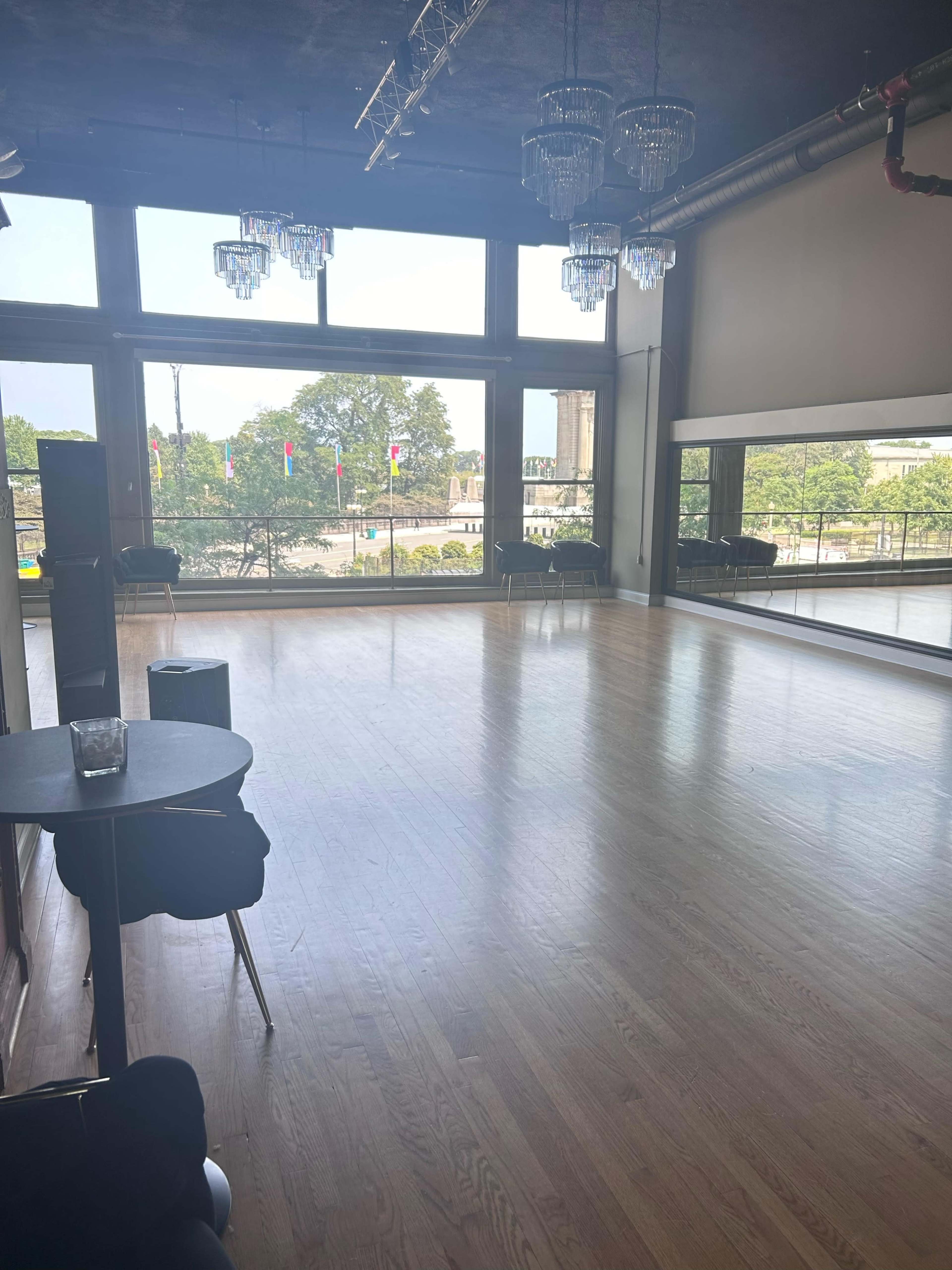 A spacious indoor dance studio features large windows with a view of a park and cityscape, illuminated by natural light.