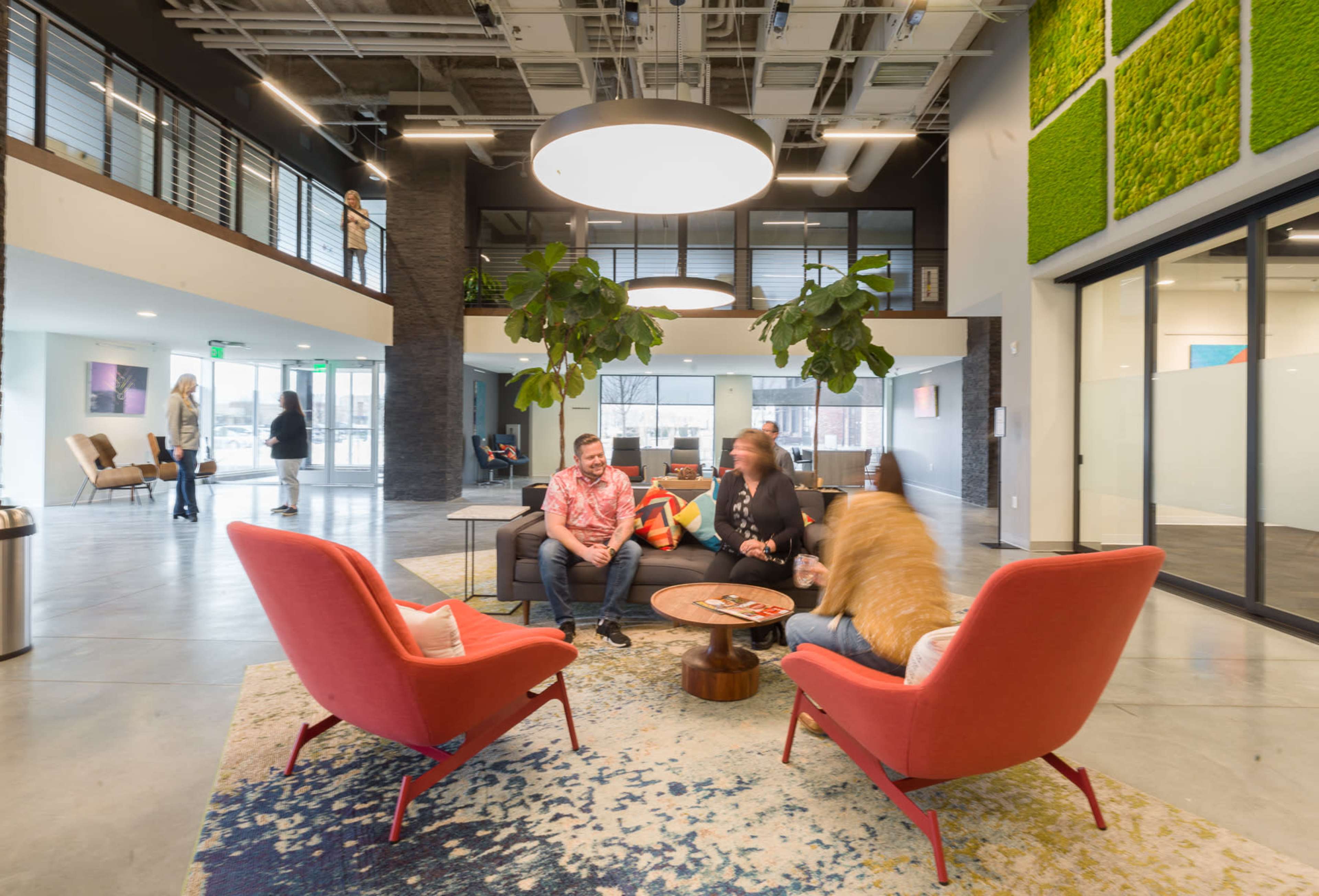 The image shows a modern office lobby with red chairs, a coffee table, greenery, and people interacting in a spacious, well-lit area.