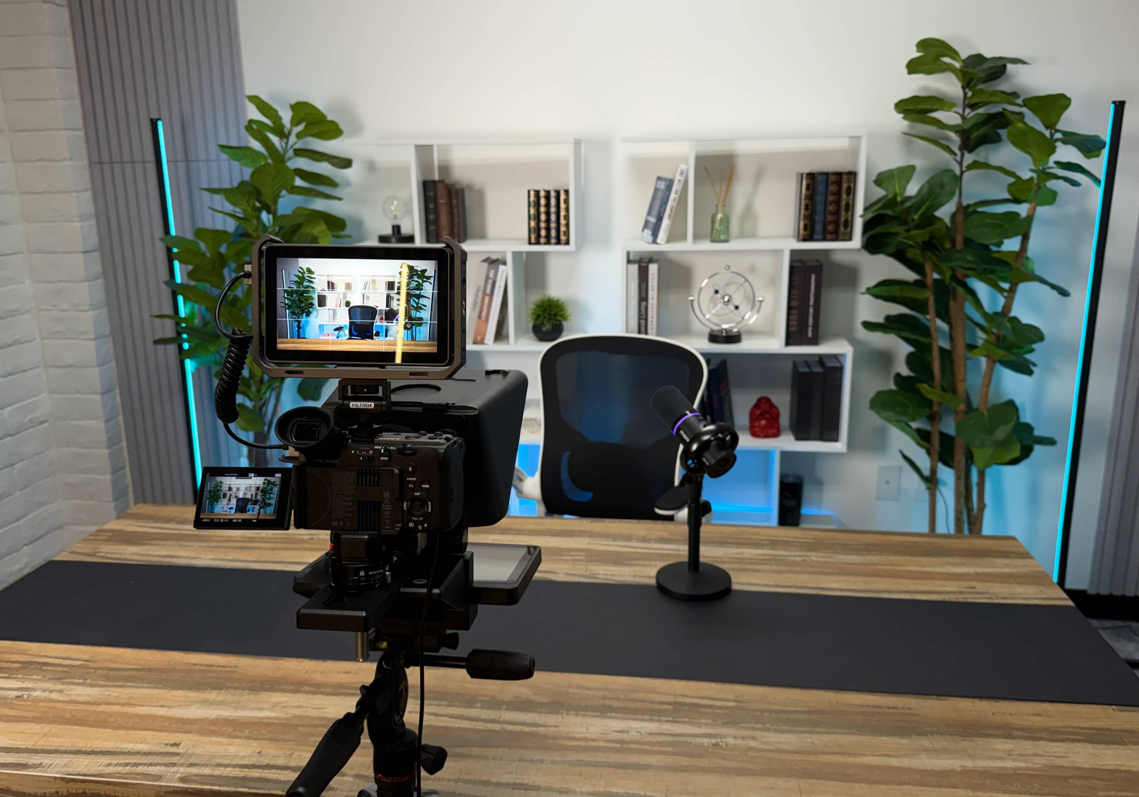 A camera is positioned on a tripod facing an empty office desk with a microphone and a background of shelves decorated with books and plants.