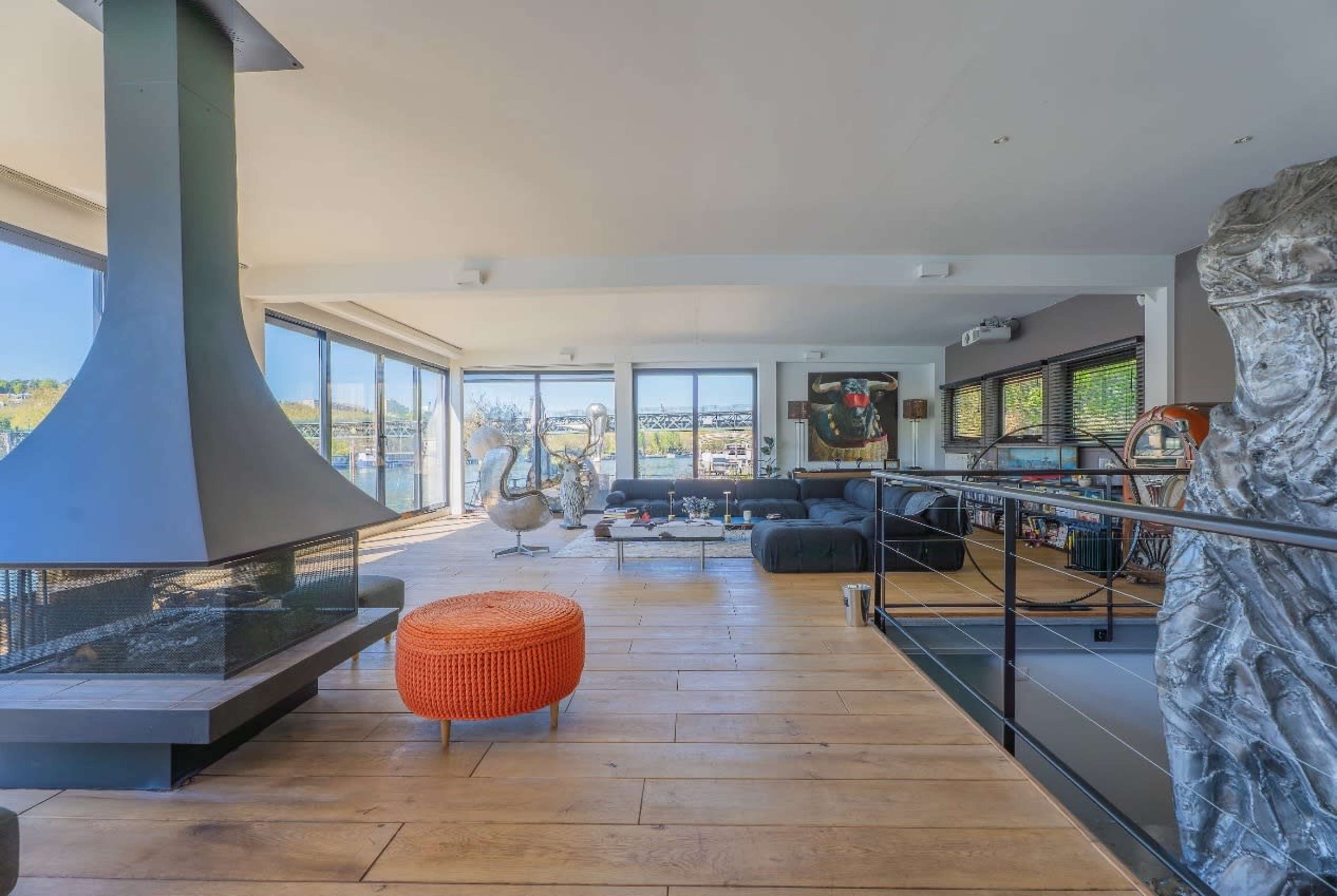 The image shows a modern living room with large windows, a central fireplace, a round orange ottoman, and contemporary furniture.