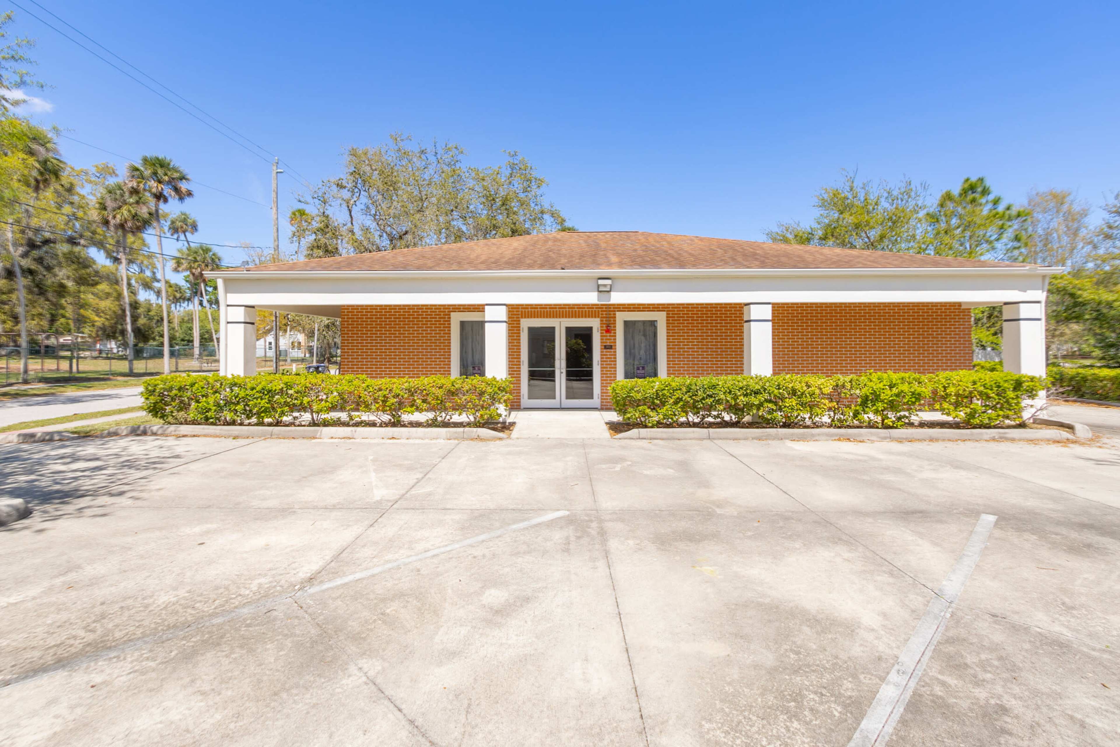 The image shows a single-story building with a brown roof and brick facade, surrounded by neatly trimmed shrubs and a concrete parking lot.