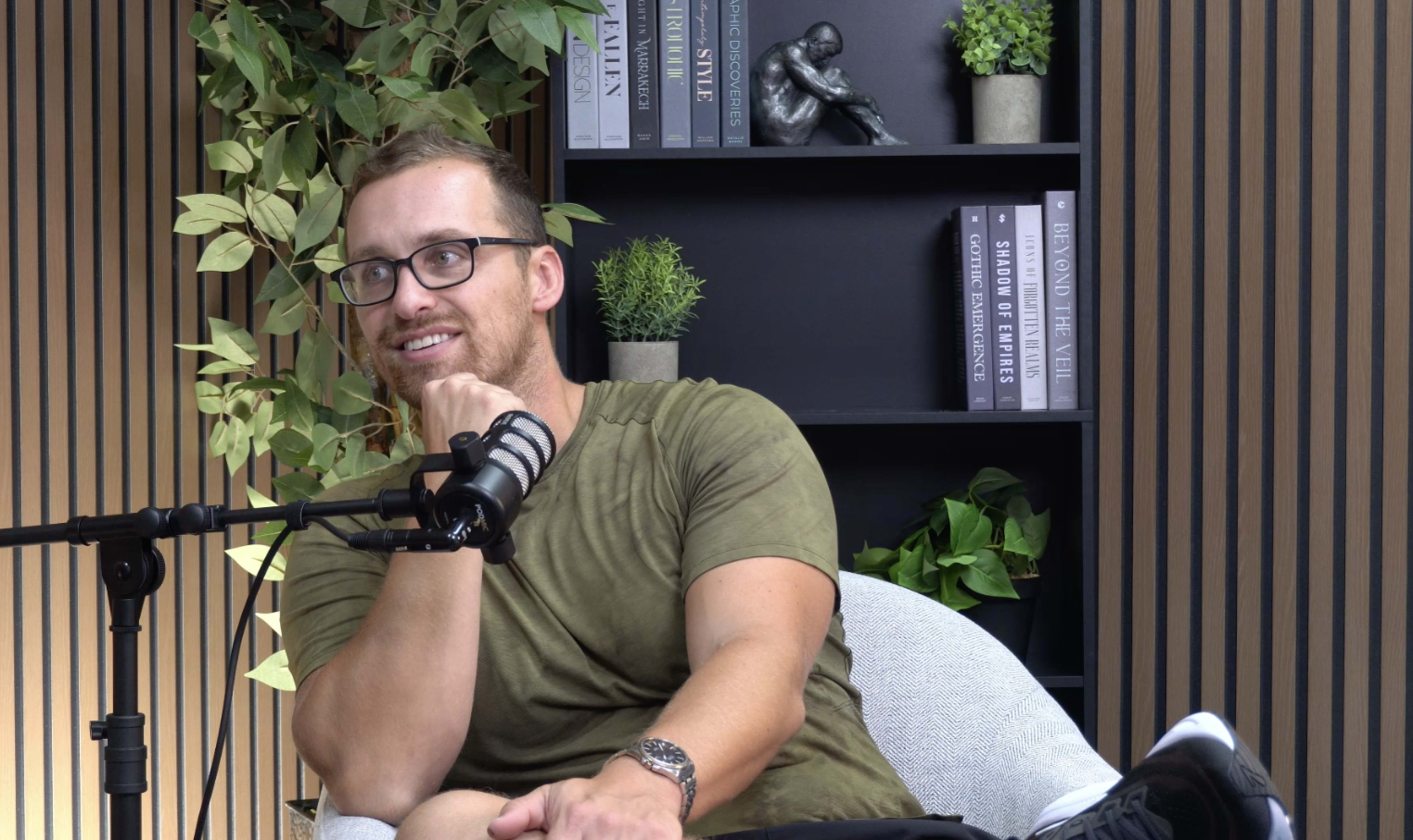 A man wearing glasses sits in a chair with a microphone nearby, in front of a bookshelf adorned with plants and decorative items.