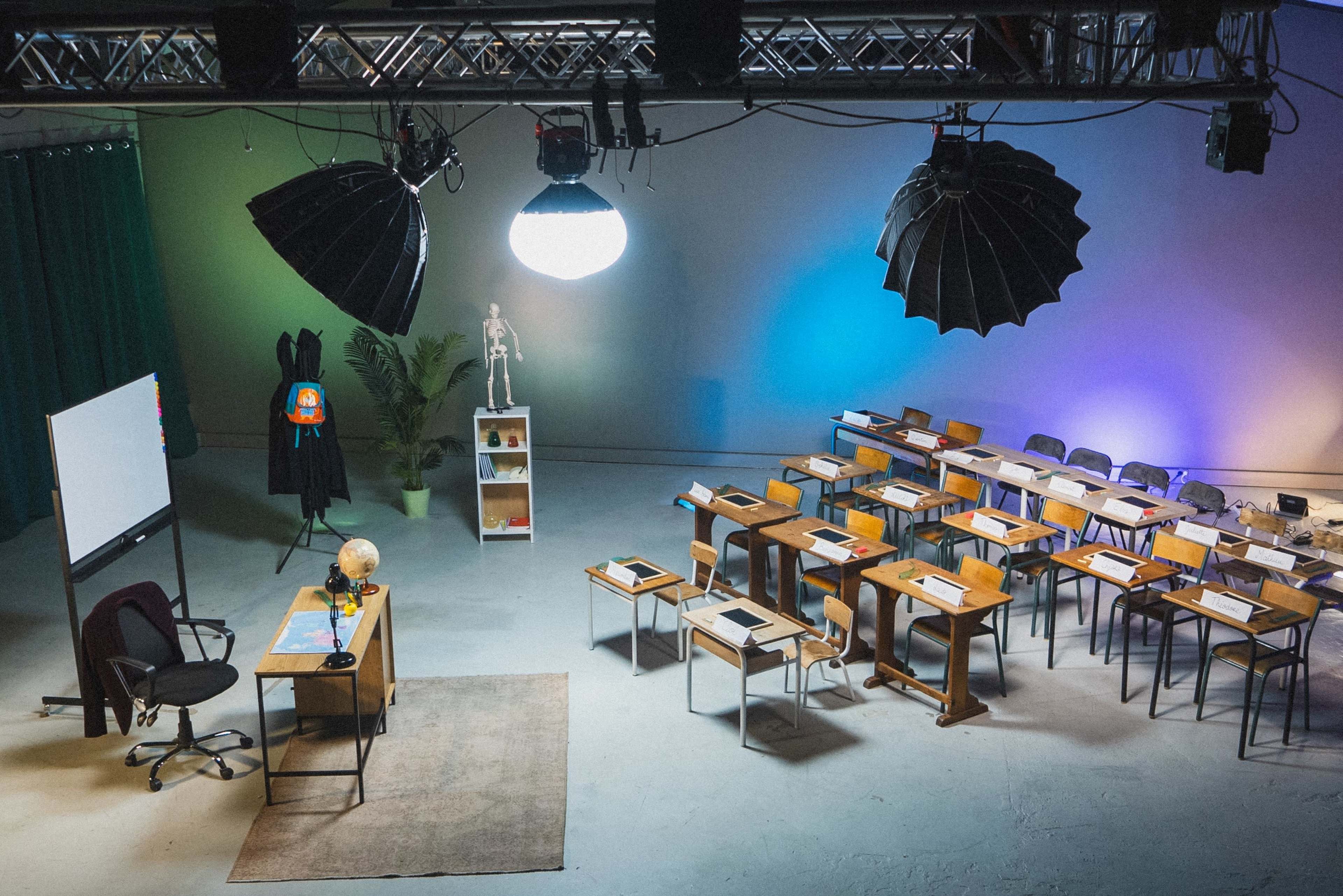 The image shows a classroom setup with wooden desks arranged in rows, a blackboard, studio lights hanging from the ceiling, and various educational materials displayed on a shelf.