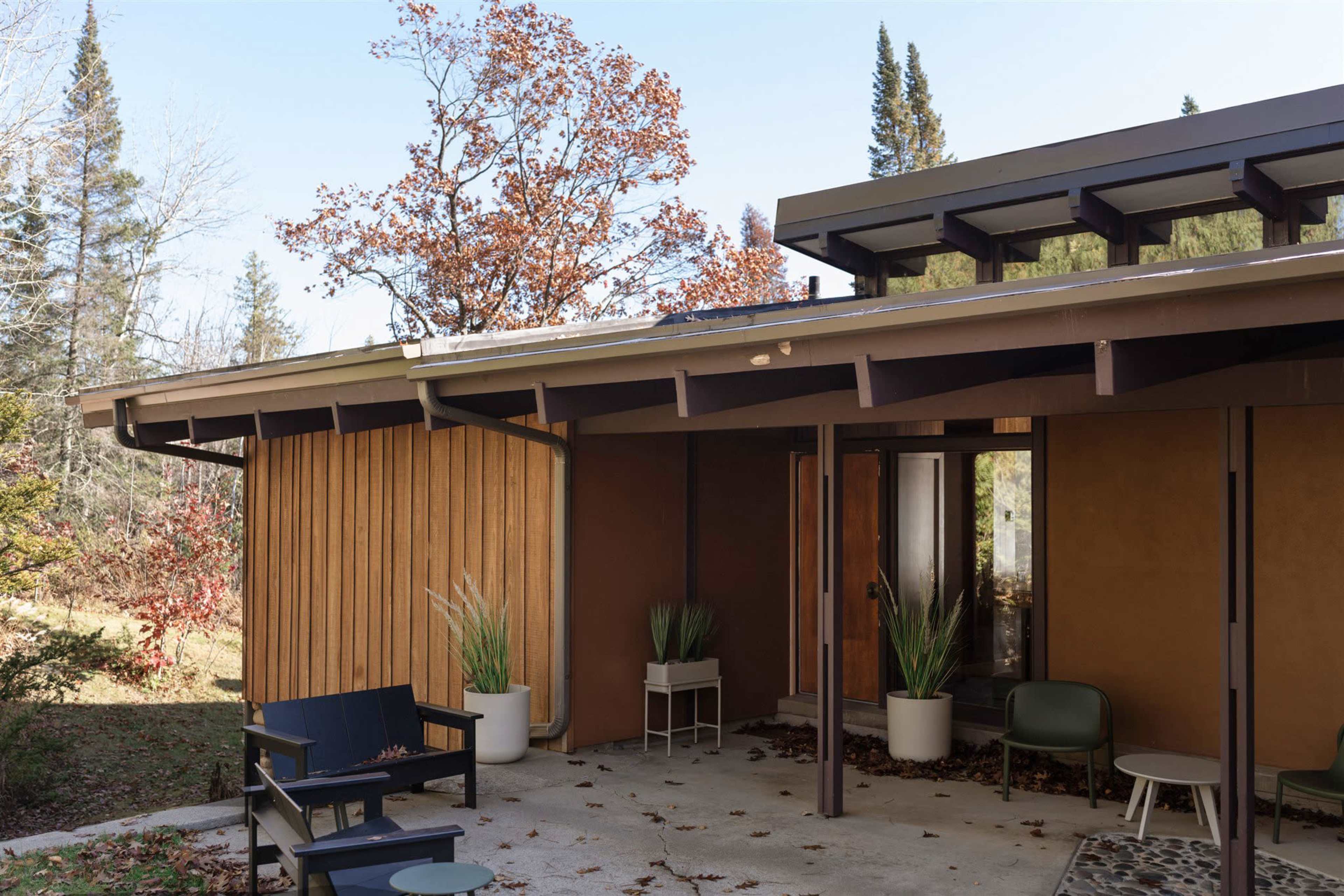 A modern home with a brown exterior features a covered patio lined with potted plants and outdoor seating, surrounded by trees.