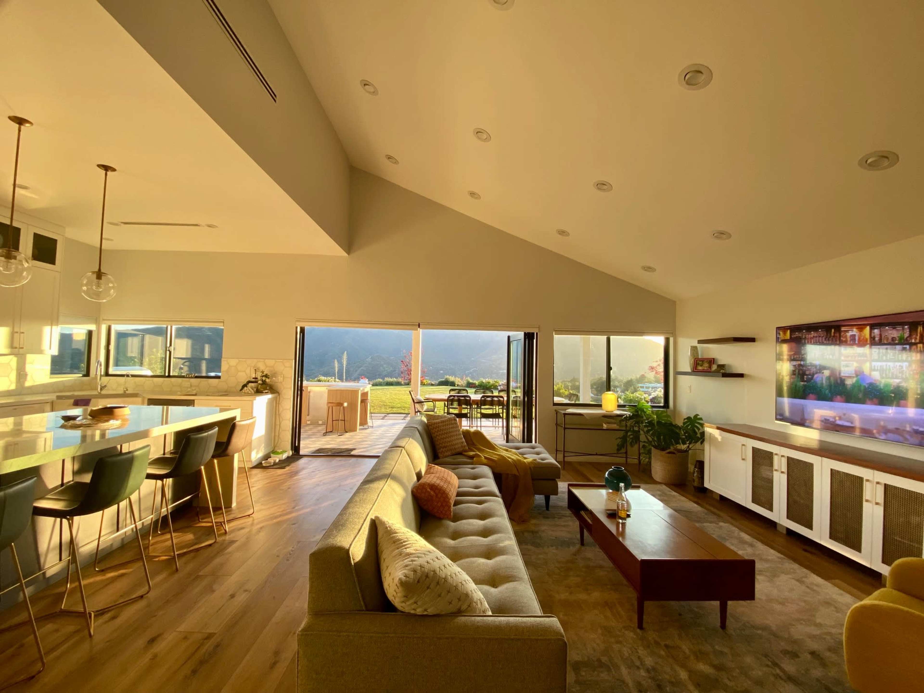The image shows a modern living room with a sectional sofa, a coffee table, and a large window opening to an outdoor patio with a mountain view.