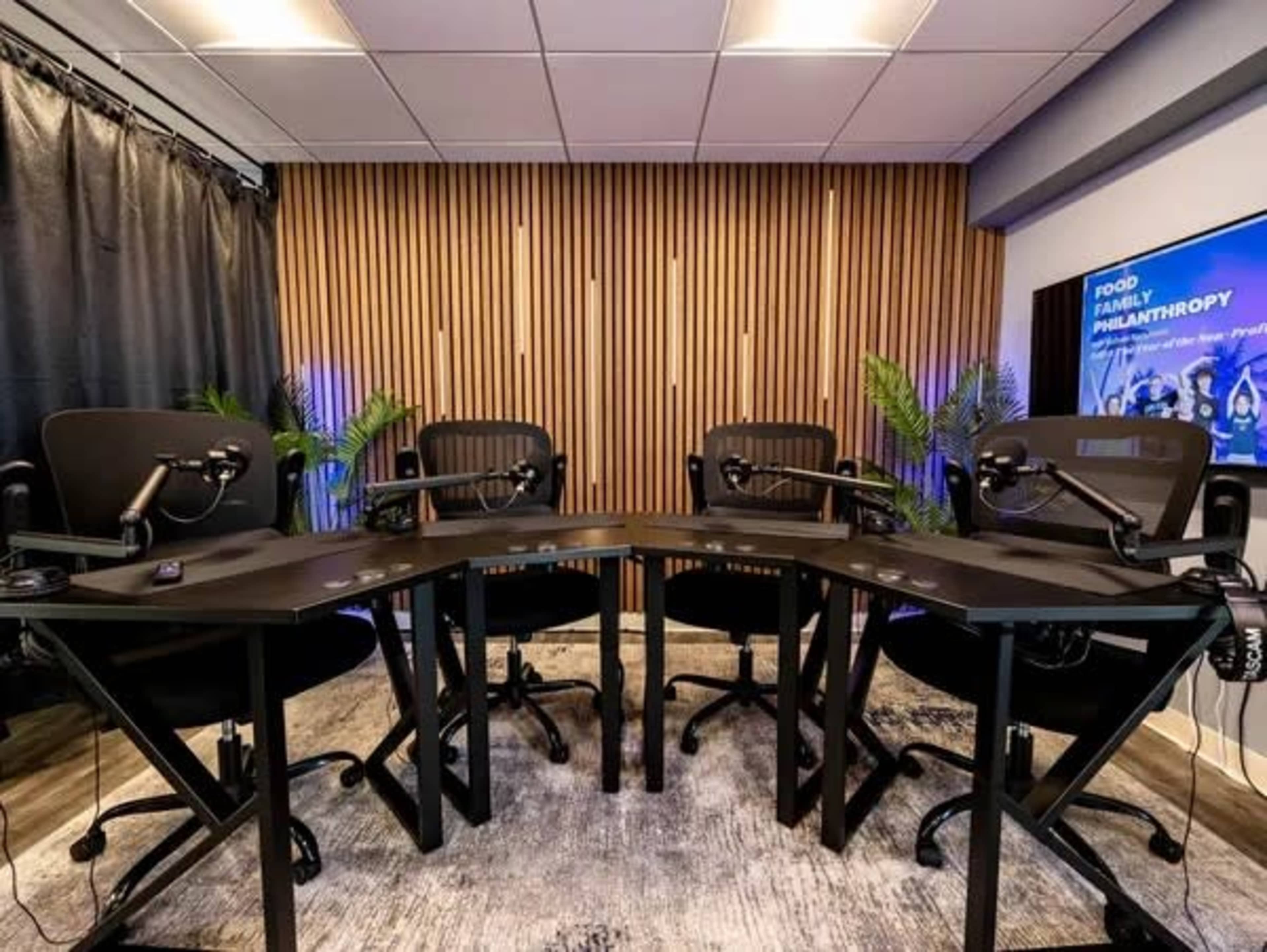 The image shows a podcast recording studio with four black chairs arranged around a hexagonal table, equipped with microphones and surrounded by decorative plants and wooden paneling.