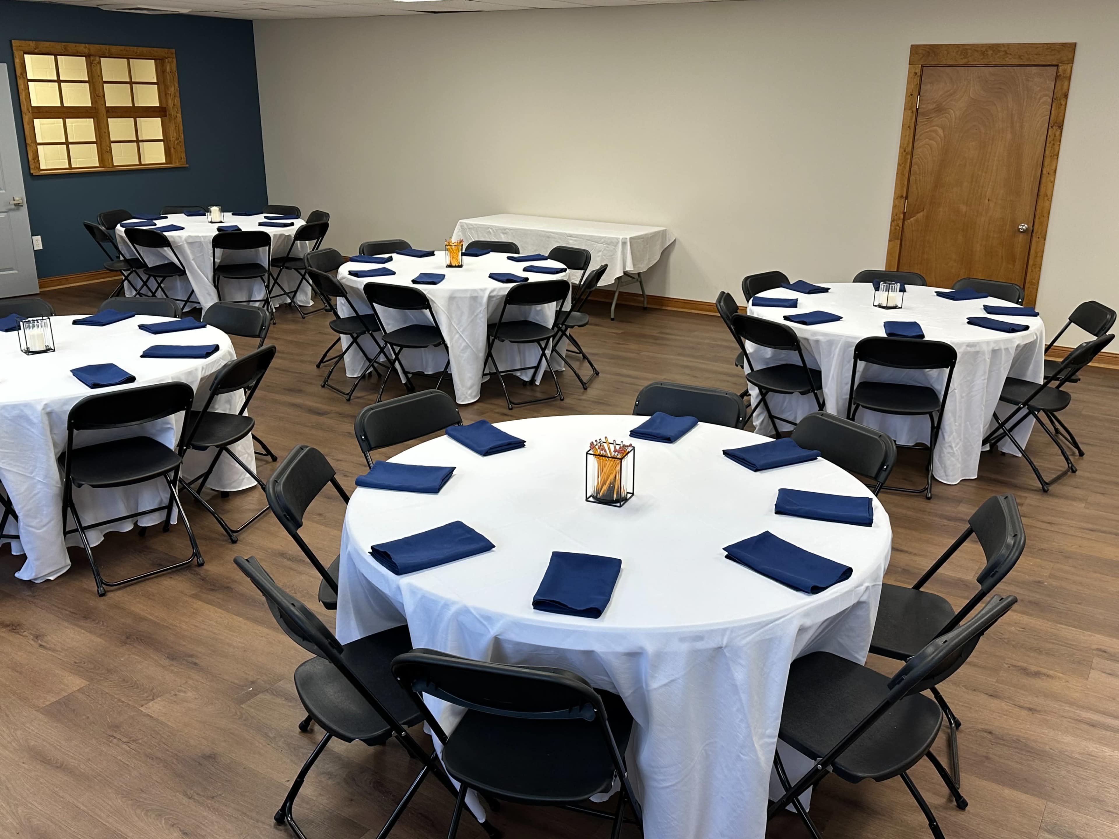 The image shows a room set up for an event with multiple round tables covered in white tablecloths and navy blue napkins, arranged with black folding chairs.