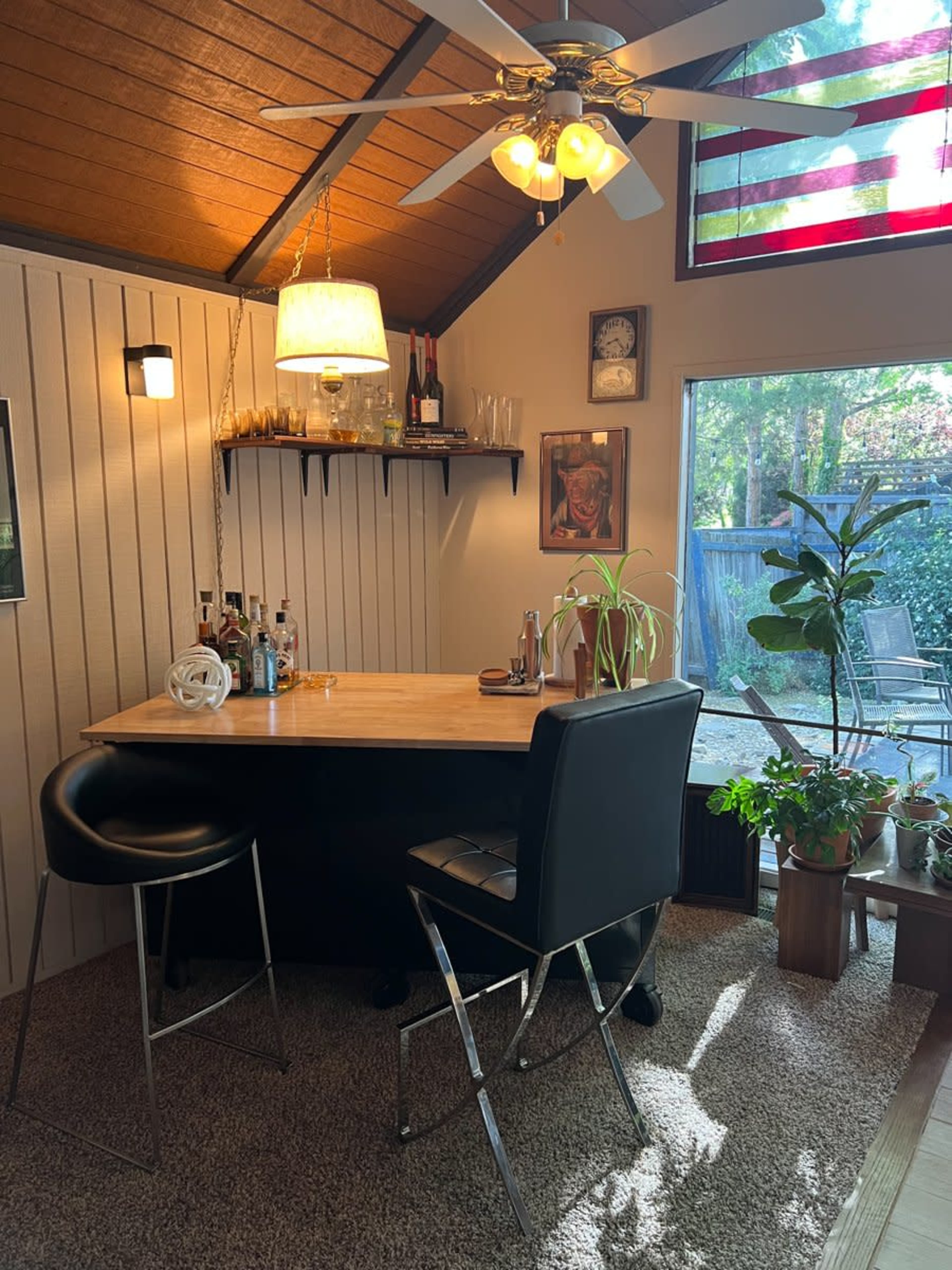A cozy home bar setup with a wooden countertop, black stools, and a wall adorned with shelves holding glasses and liquor bottles, surrounded by potted plants and a large window.