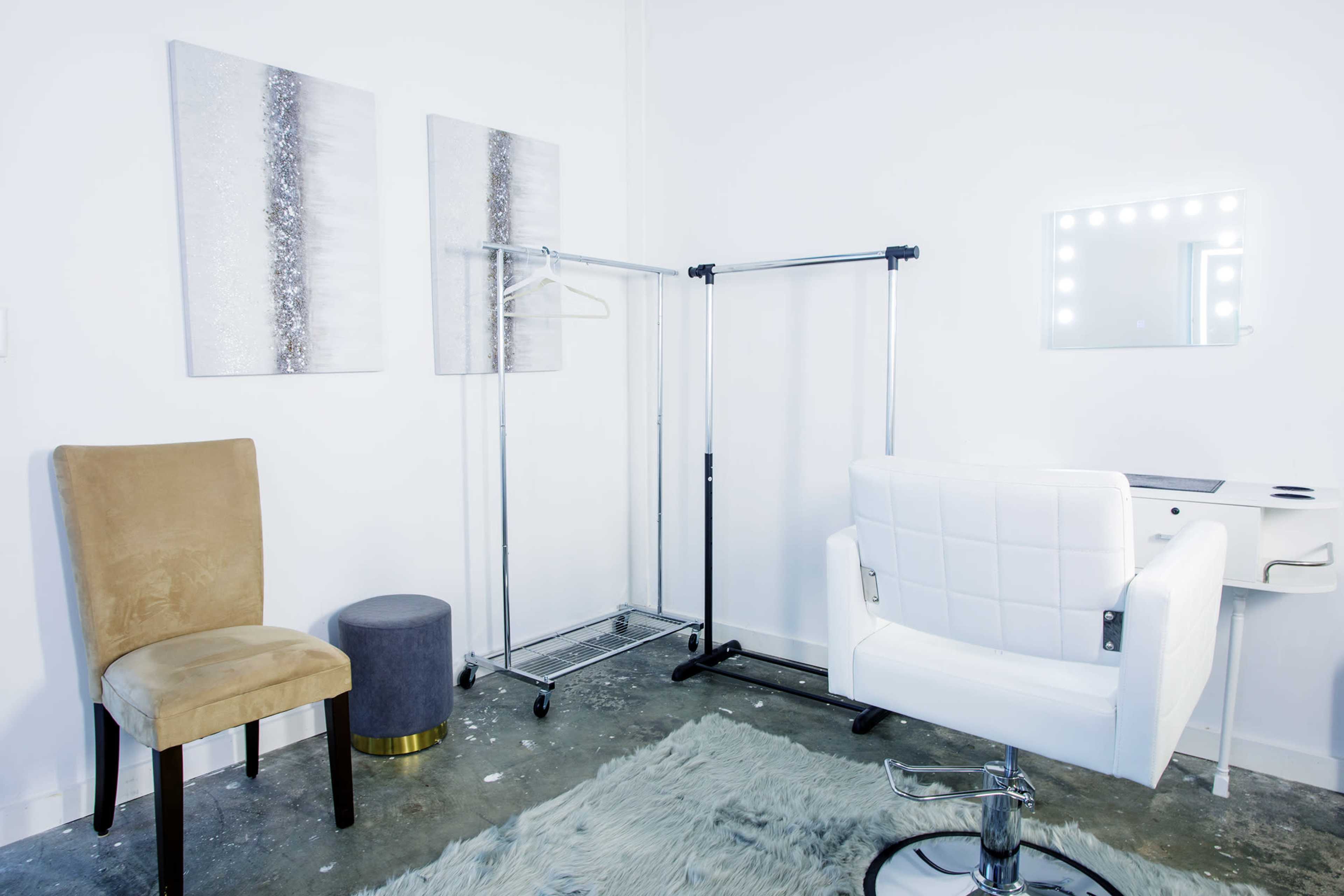 The image shows a minimalist hair salon interior featuring a white salon chair, a small stool, two wall-mounted paintings, and a clothing rack against a white wall.