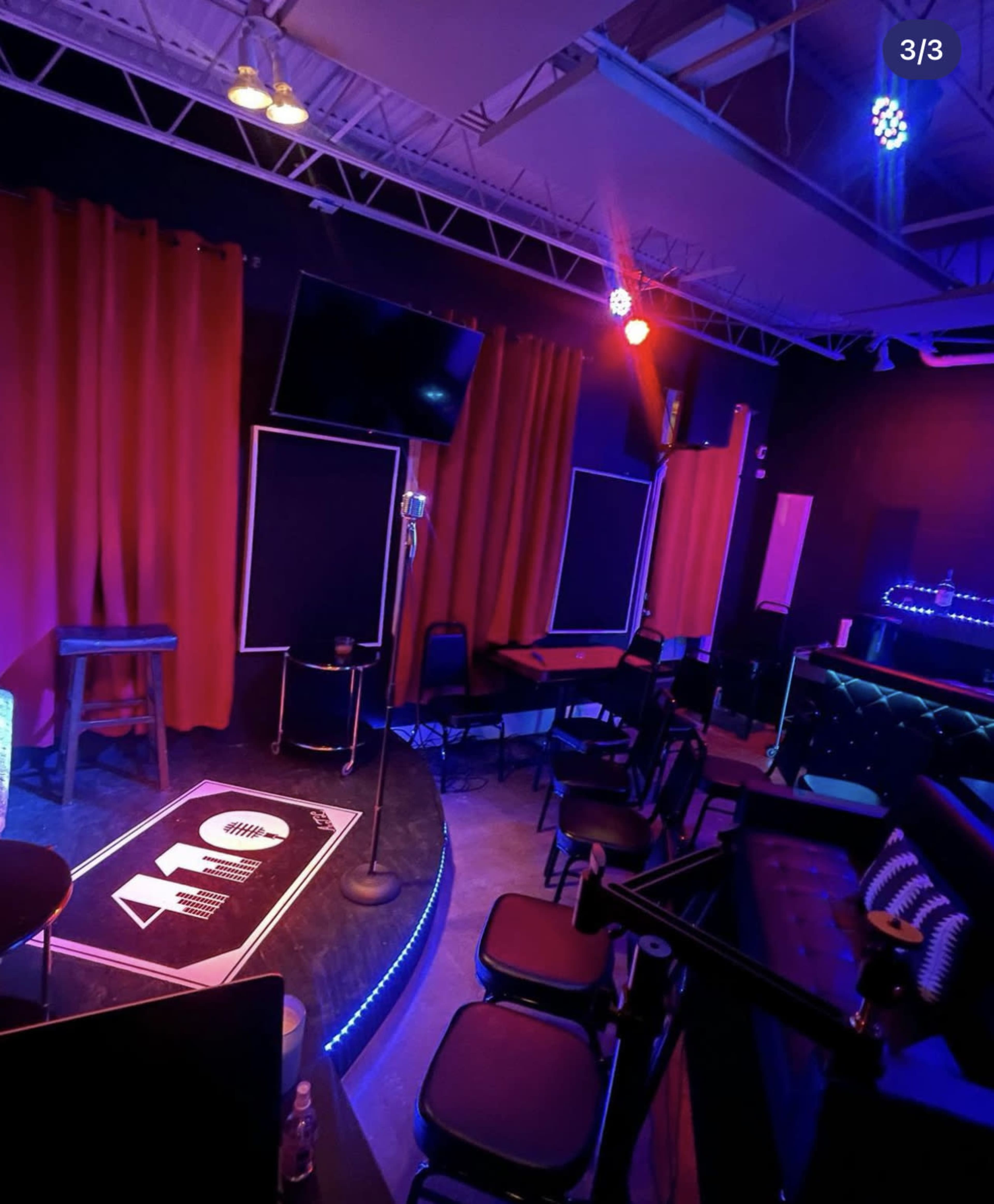 A dimly lit performance space featuring a stage with a microphone, illuminated seating areas, and red curtains.