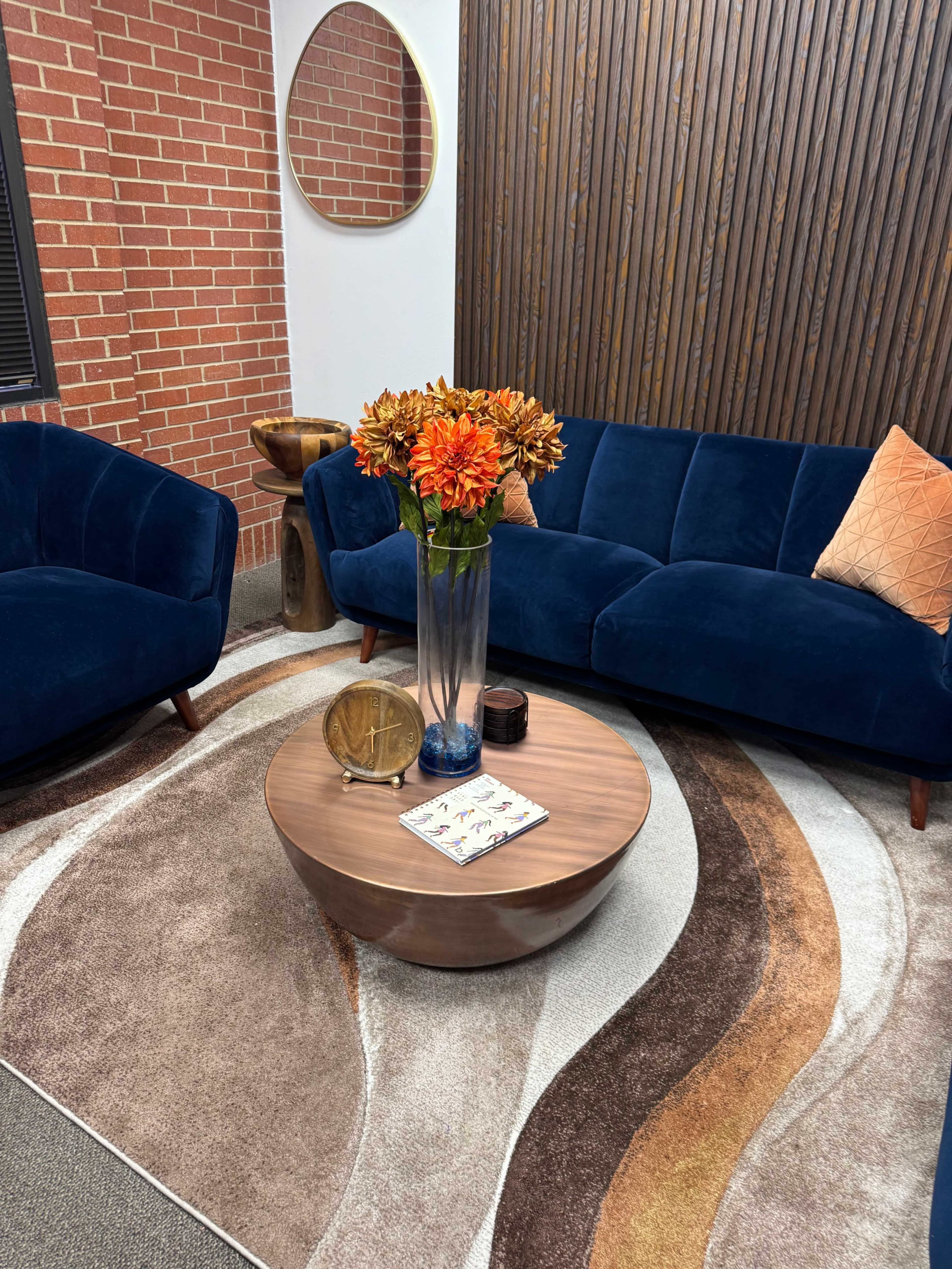 The image shows a cozy seating area featuring two blue velvet chairs and a matching sofa, a round wooden coffee table, and a tall vase with orange flowers, all set against a backdrop of wooden paneling and a brick wall.