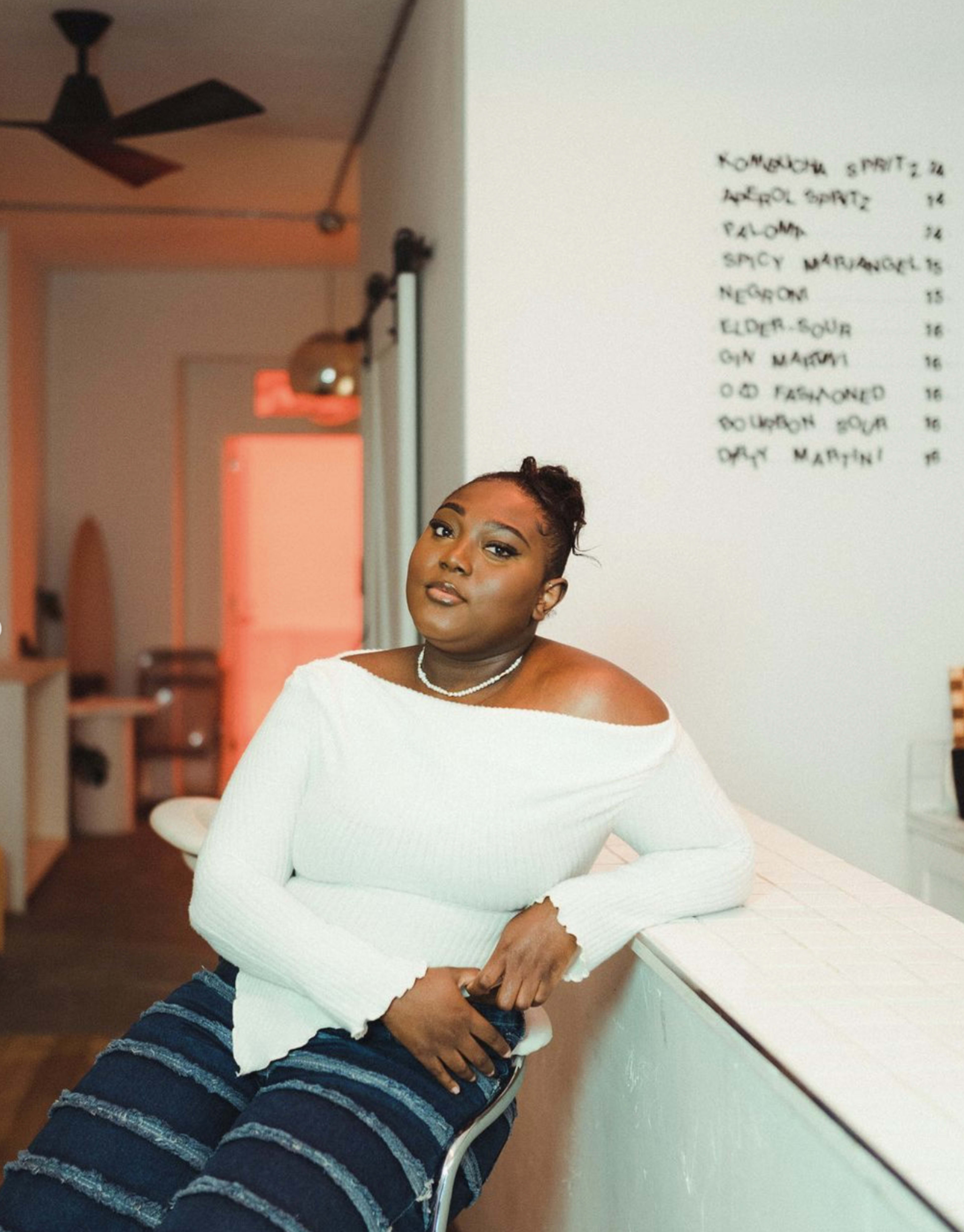 A woman sits on a barstool at a counter with a black menu on the wall behind her.