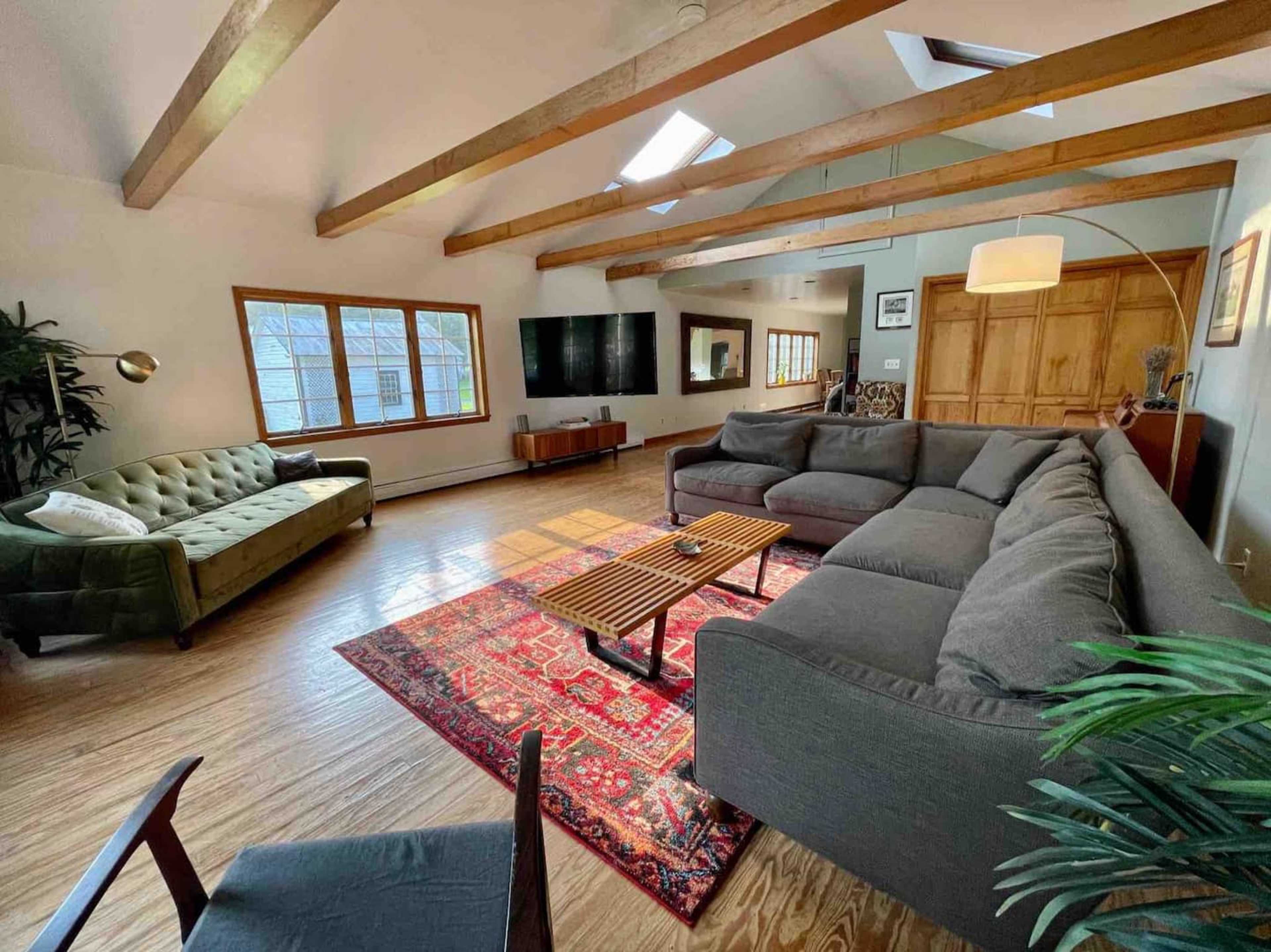 The living room features a sectional sofa, a green couch, a patterned rug, wooden beams, and a large window.