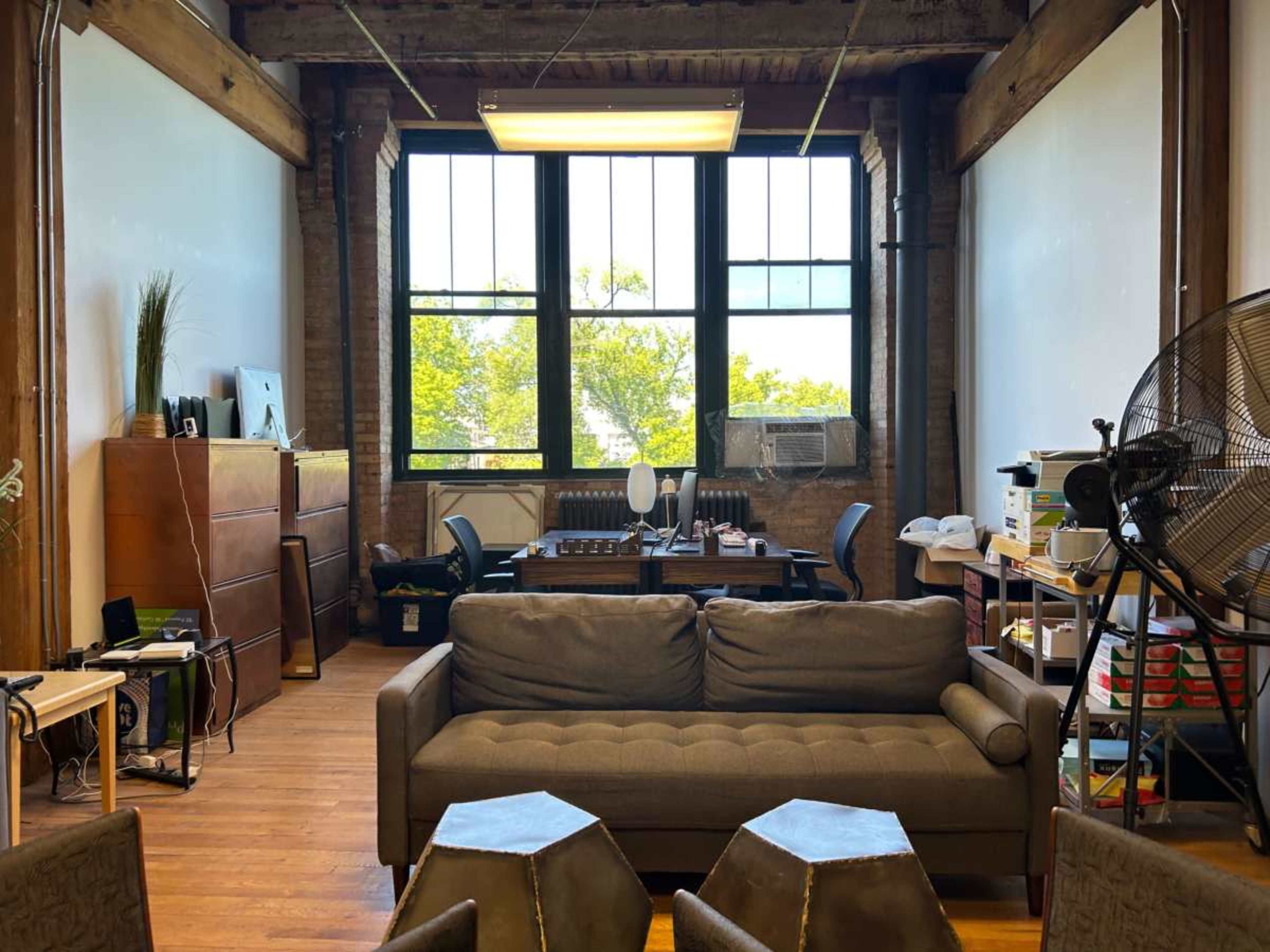The image shows a spacious office or workspace with large windows, a brown sofa in the foreground, and various desks and chairs arranged in the background.