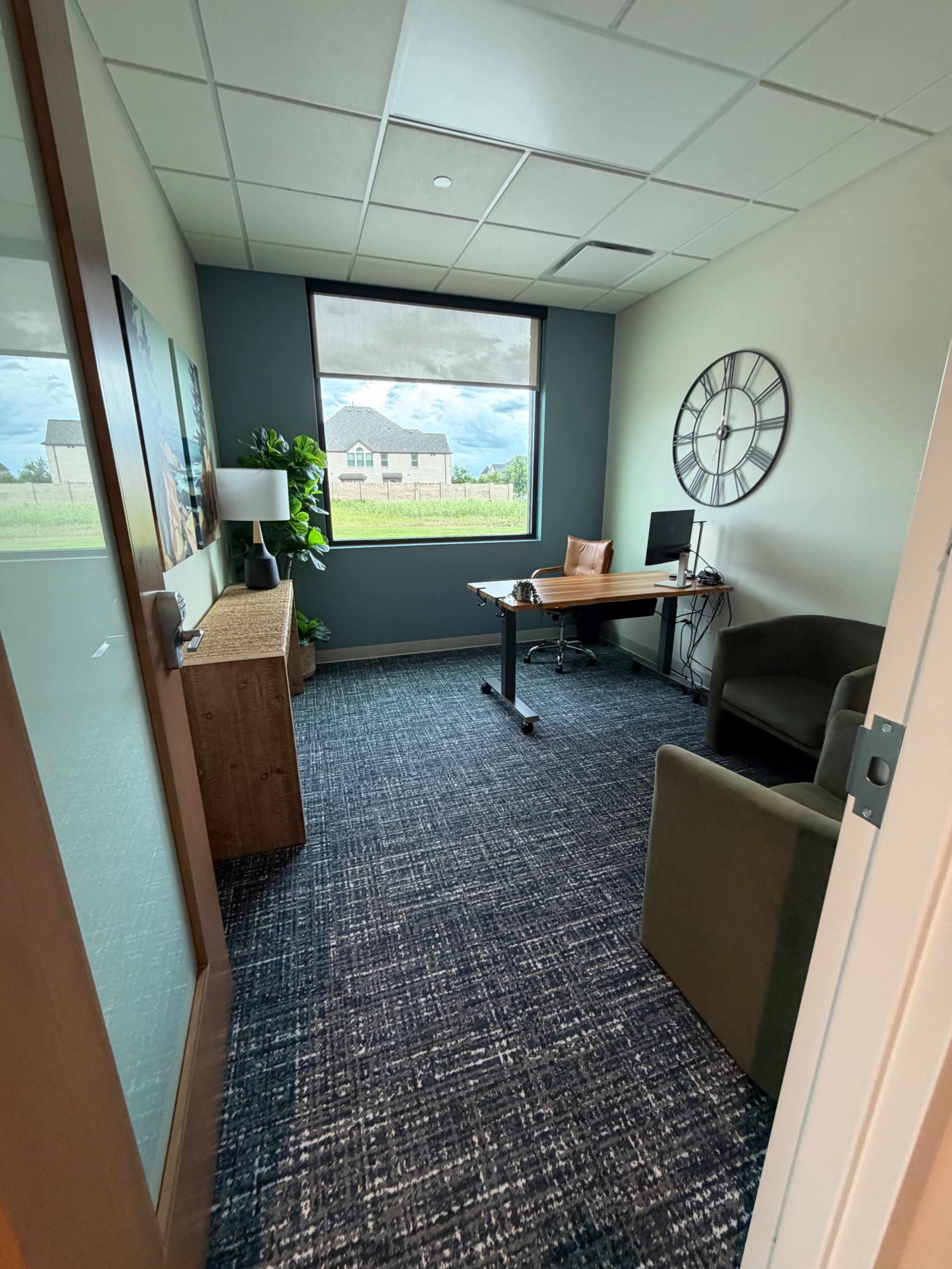 The image shows a small office space featuring a desk, two chairs, a plant, and a large window with a view of a field and a distant house.