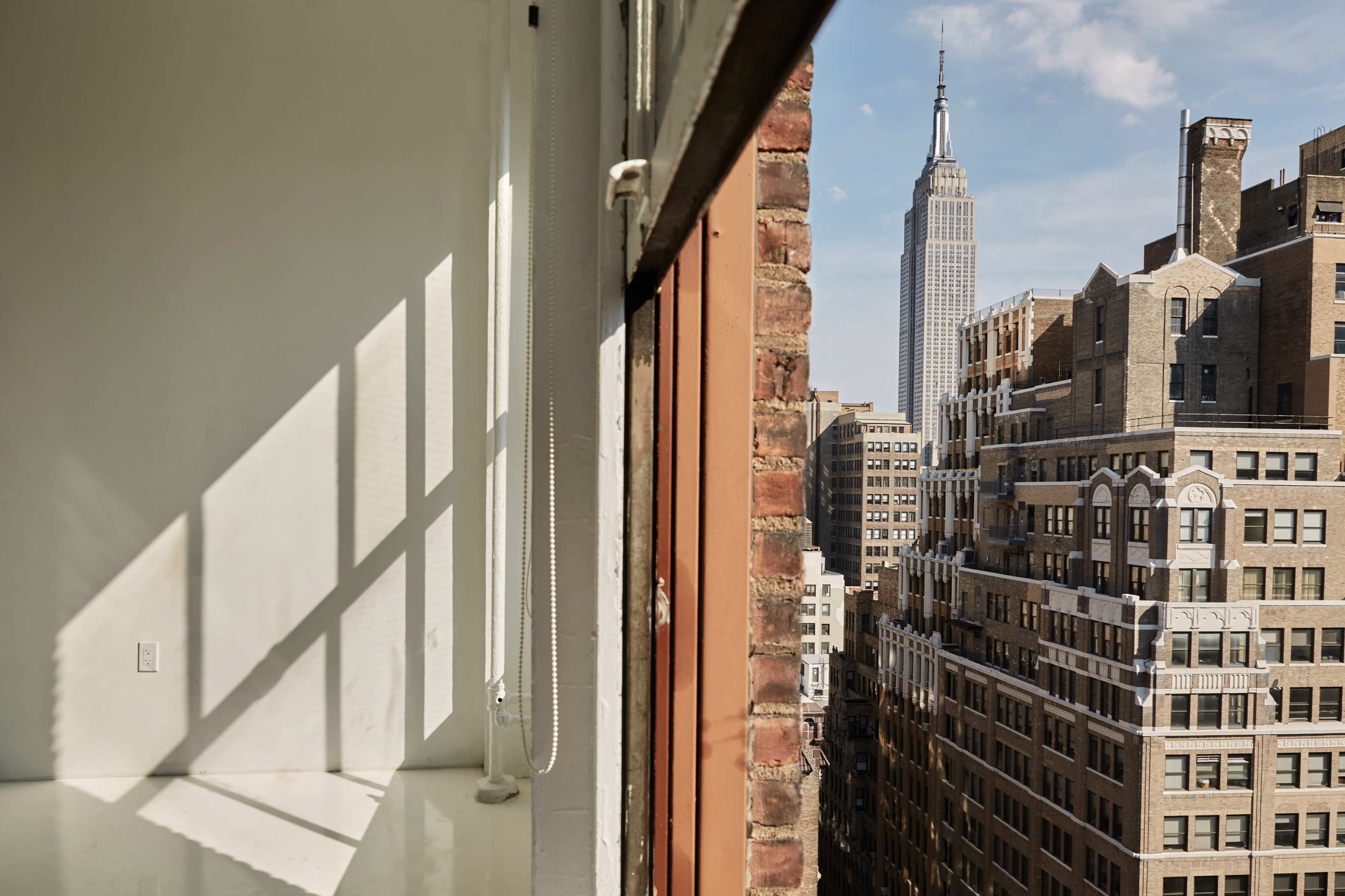 The image shows a vacant room with large windows overlooking a cityscape that includes the Empire State Building.