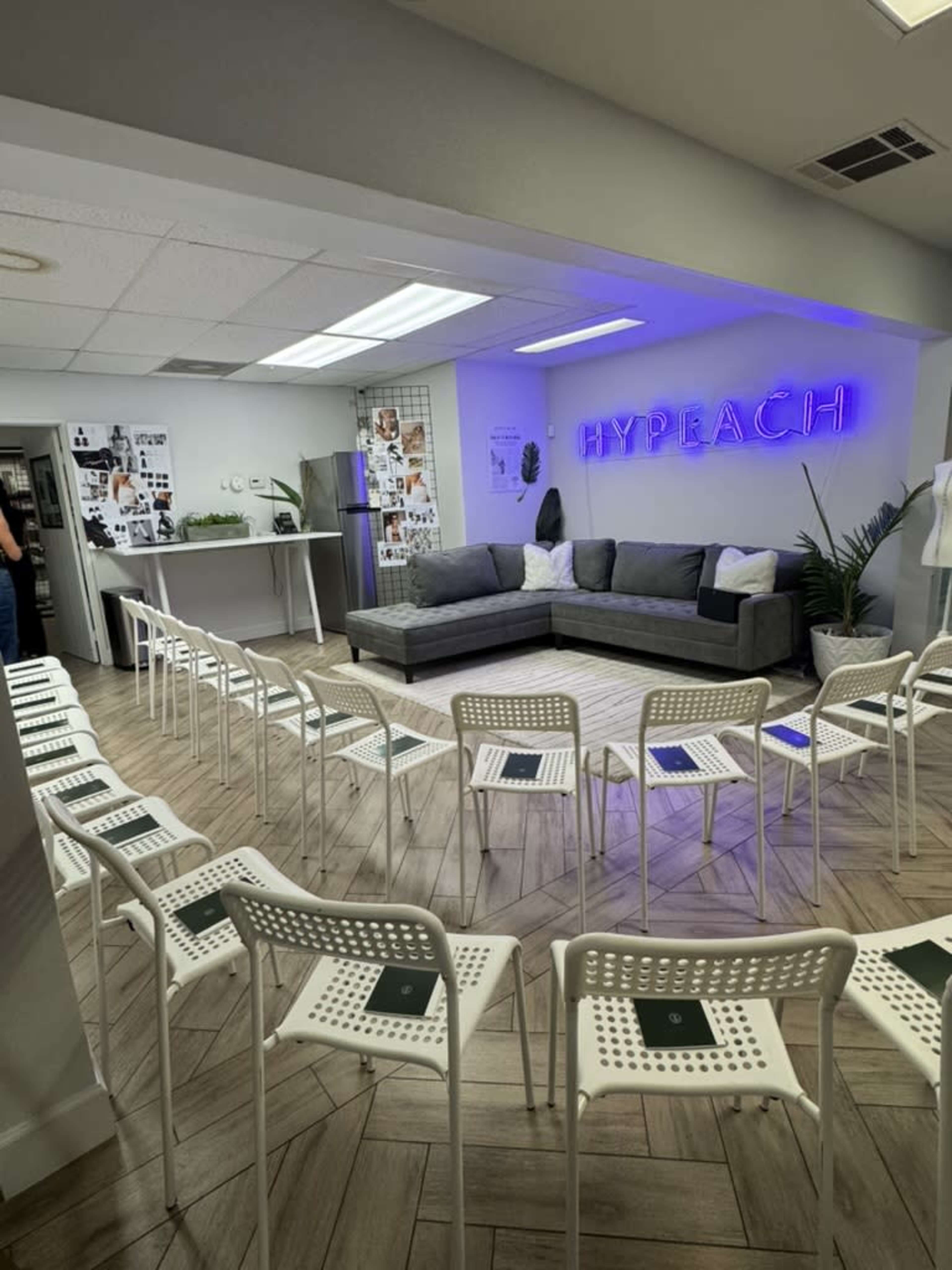 The image shows a modern room set up with a gray sectional sofa, a neon sign that reads "HYPERACH," and white chairs arranged in rows, suitable for a meeting or event.
