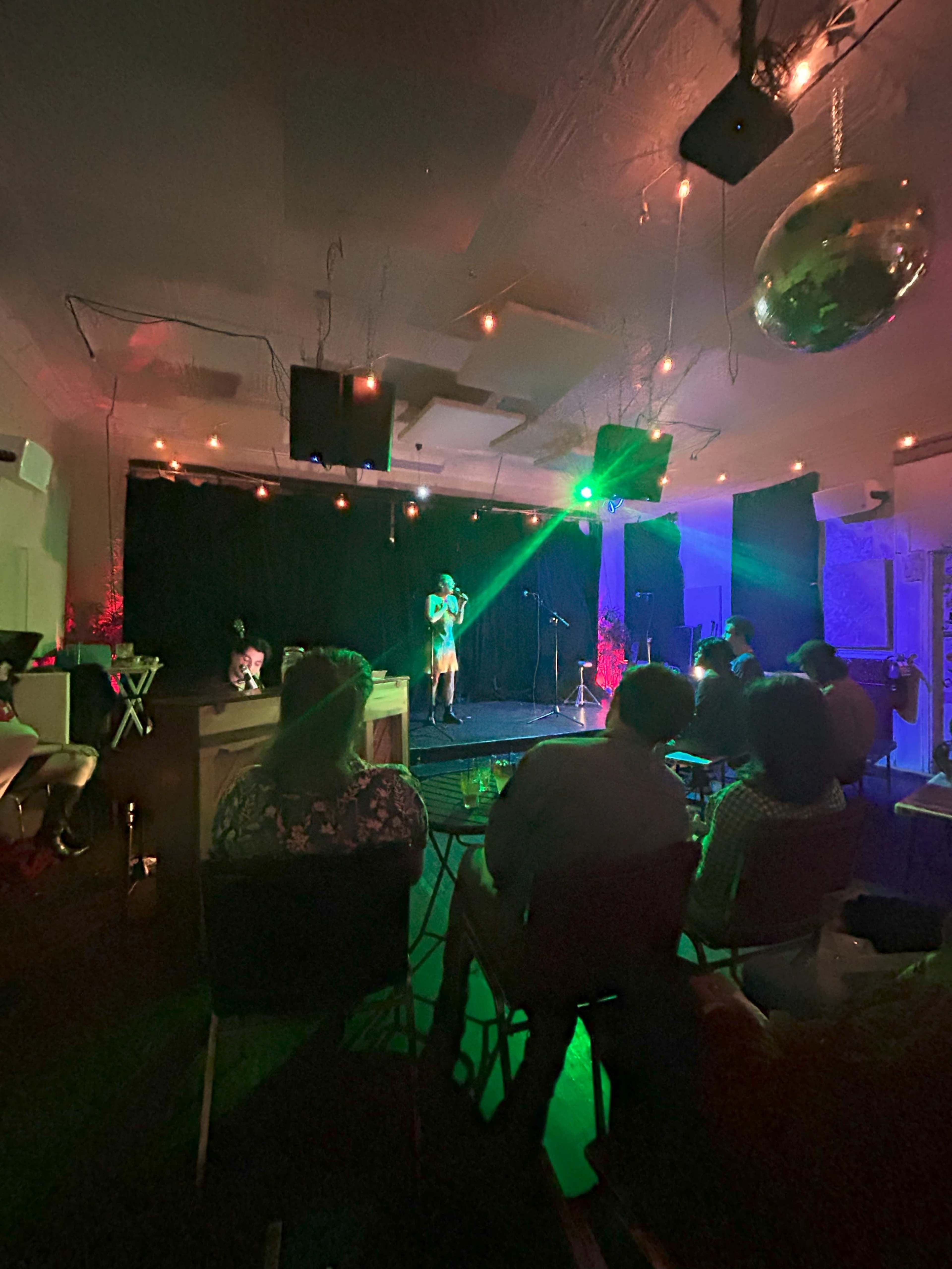 A performer stands on a stage under colorful lights while an audience watches from seated areas in a dimly lit venue.