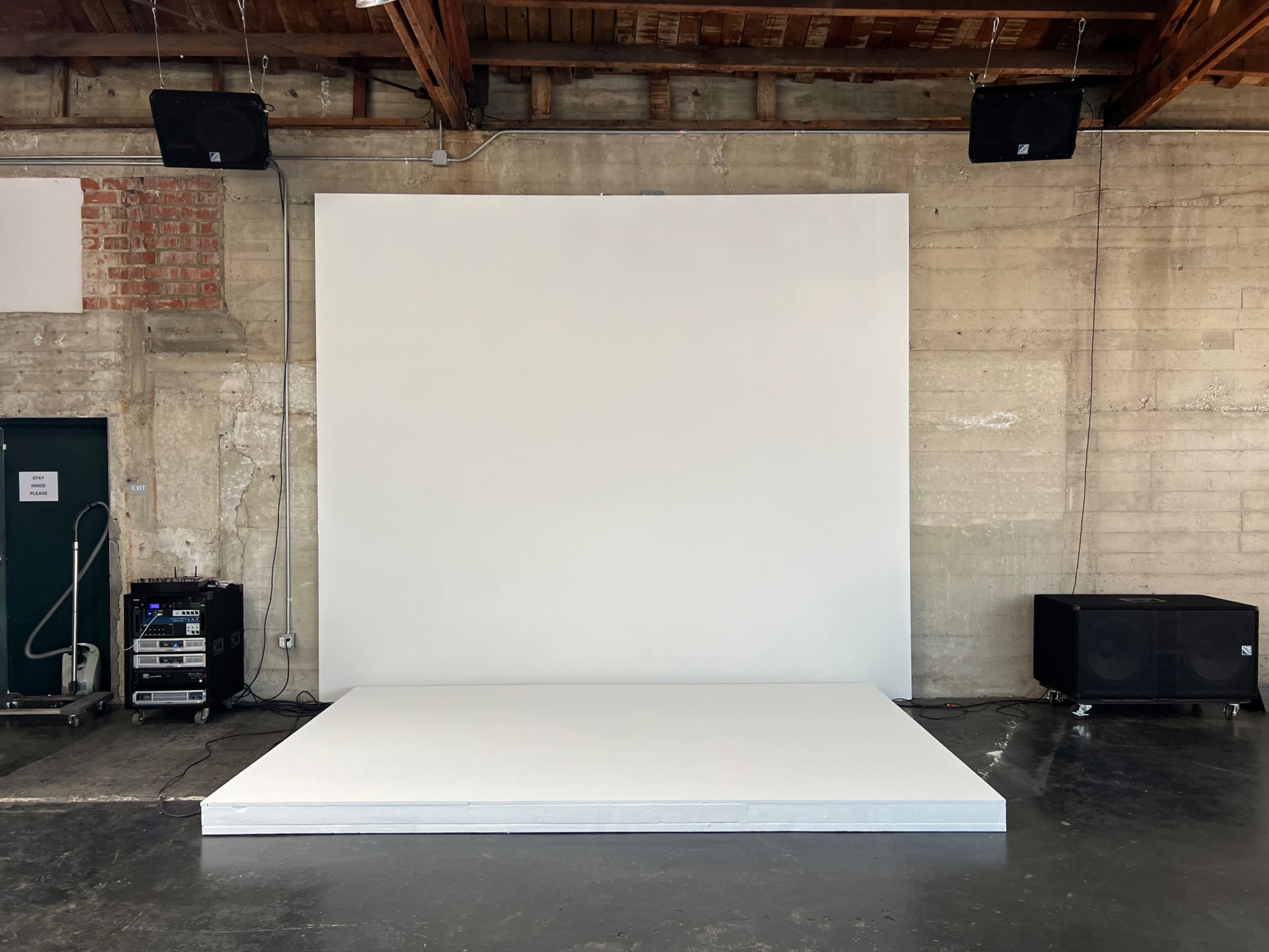 A minimalistic stage setup with a large white backdrop and a raised white platform in an industrial space.