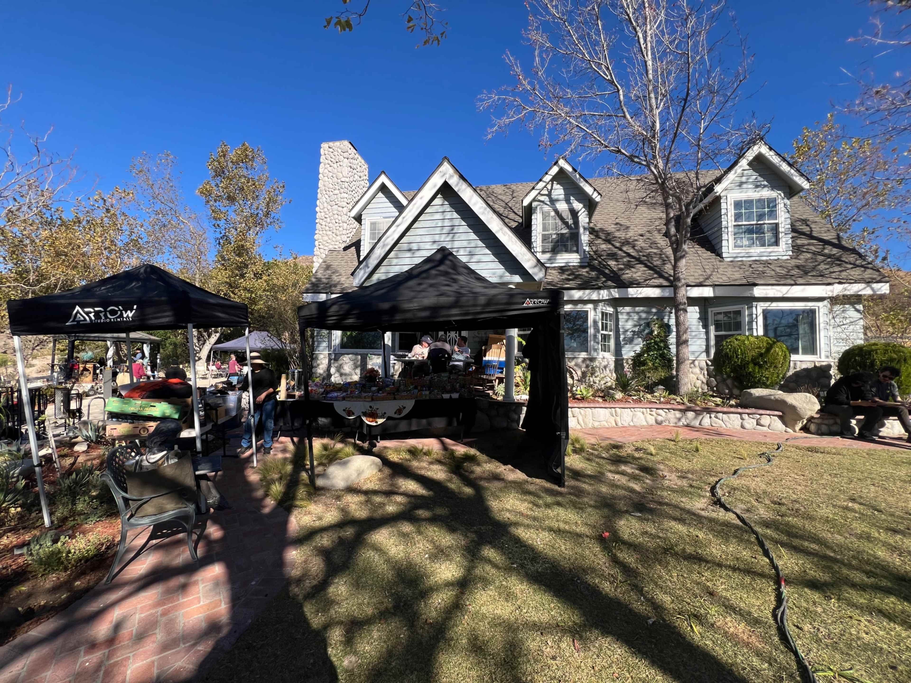 A residential house with a gray exterior is surrounded by tents set up for an outdoor market or event on a sunny day.