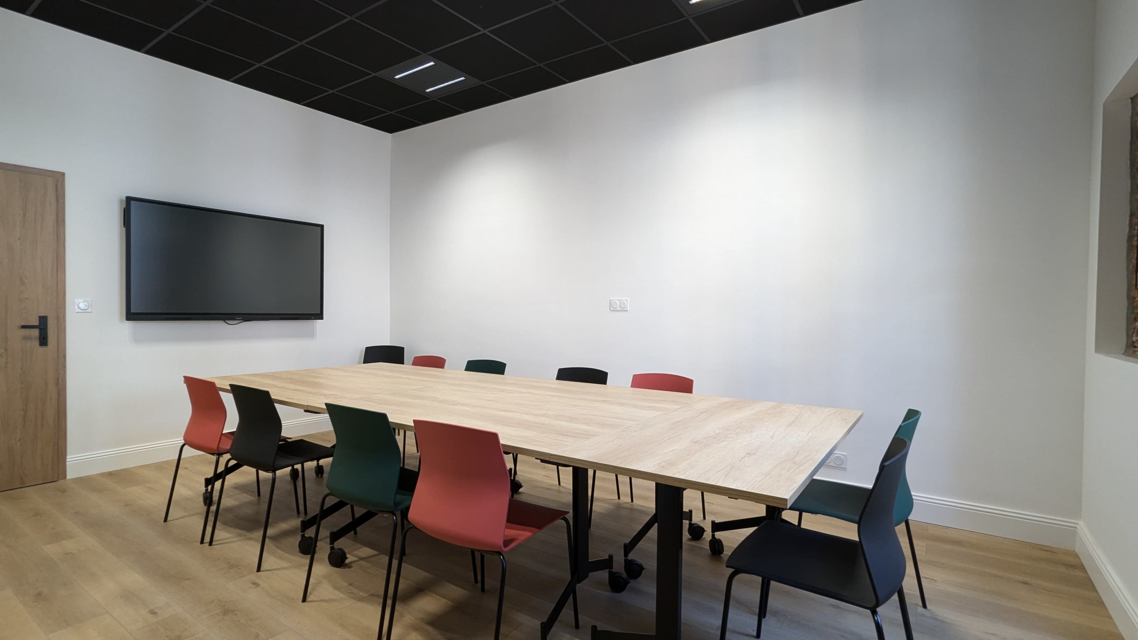 A spacious conference room features a large wooden table surrounded by colorful chairs and a wall-mounted screen.