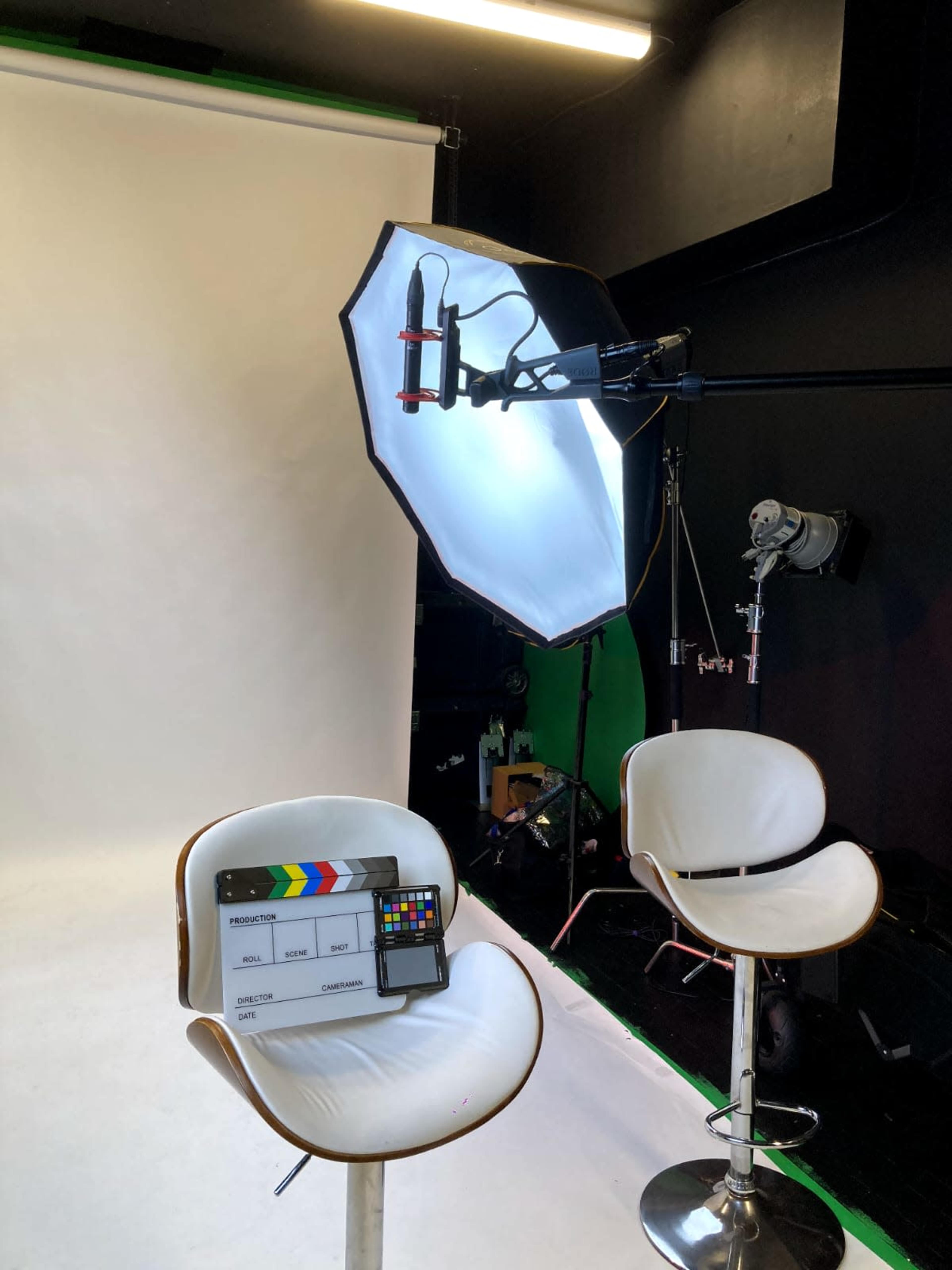 The image shows a photography or video studio setup featuring two white chairs, a light source with a softbox, and a clapperboard placed on a table.