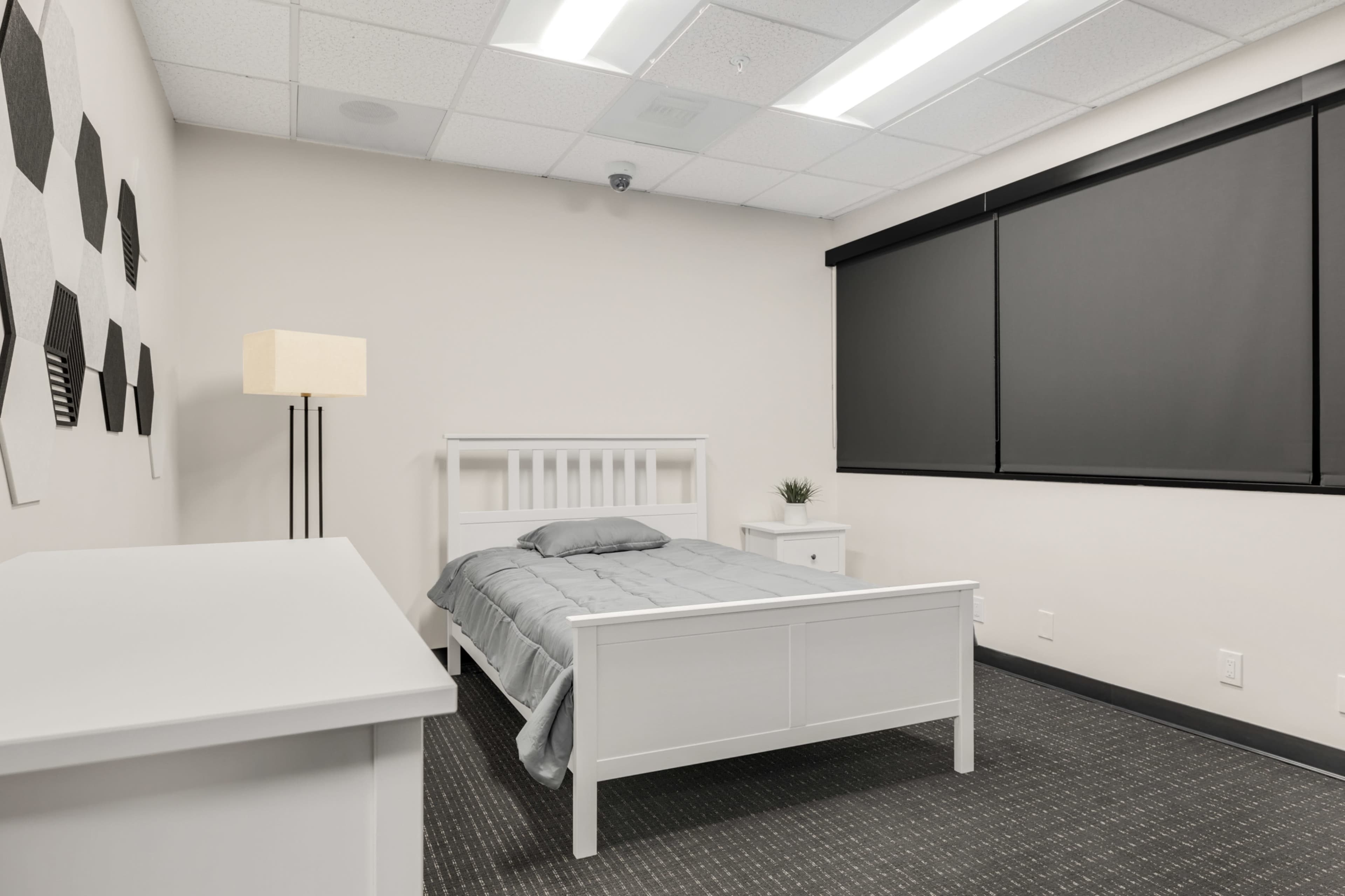 The image shows a minimalist bedroom with a white bed, bedside table, a lamp, and a dark window covering.