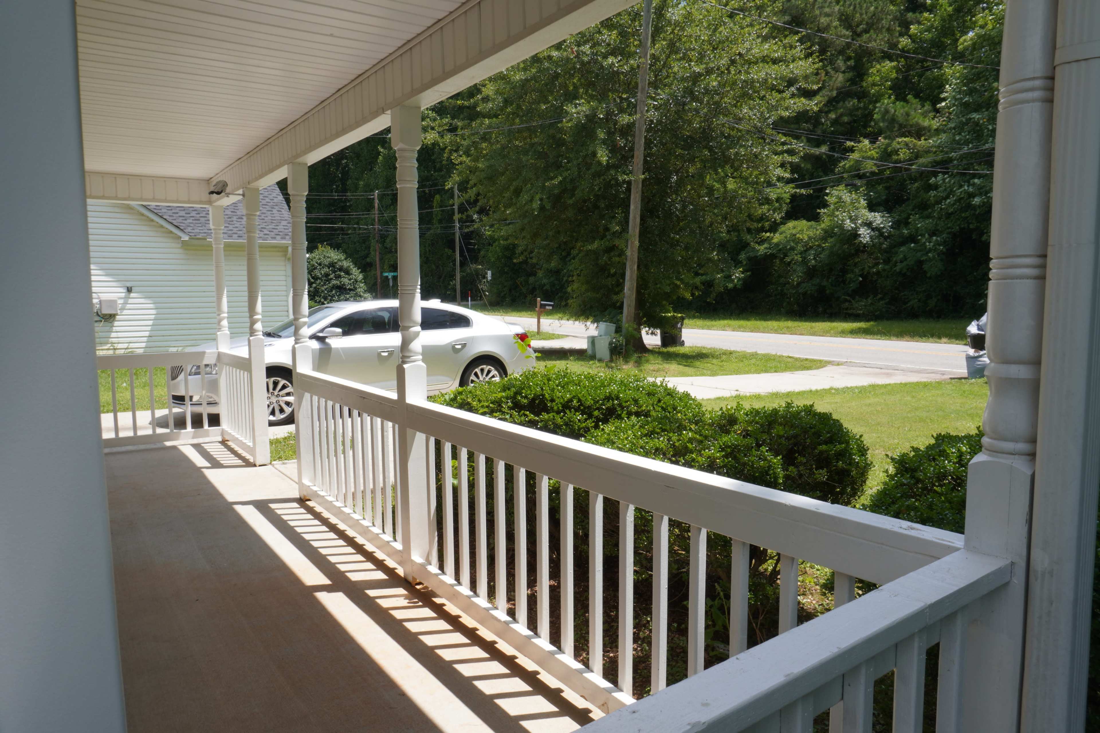 A porch with white railings overlooking a grassy area and a road, with cars parked nearby.