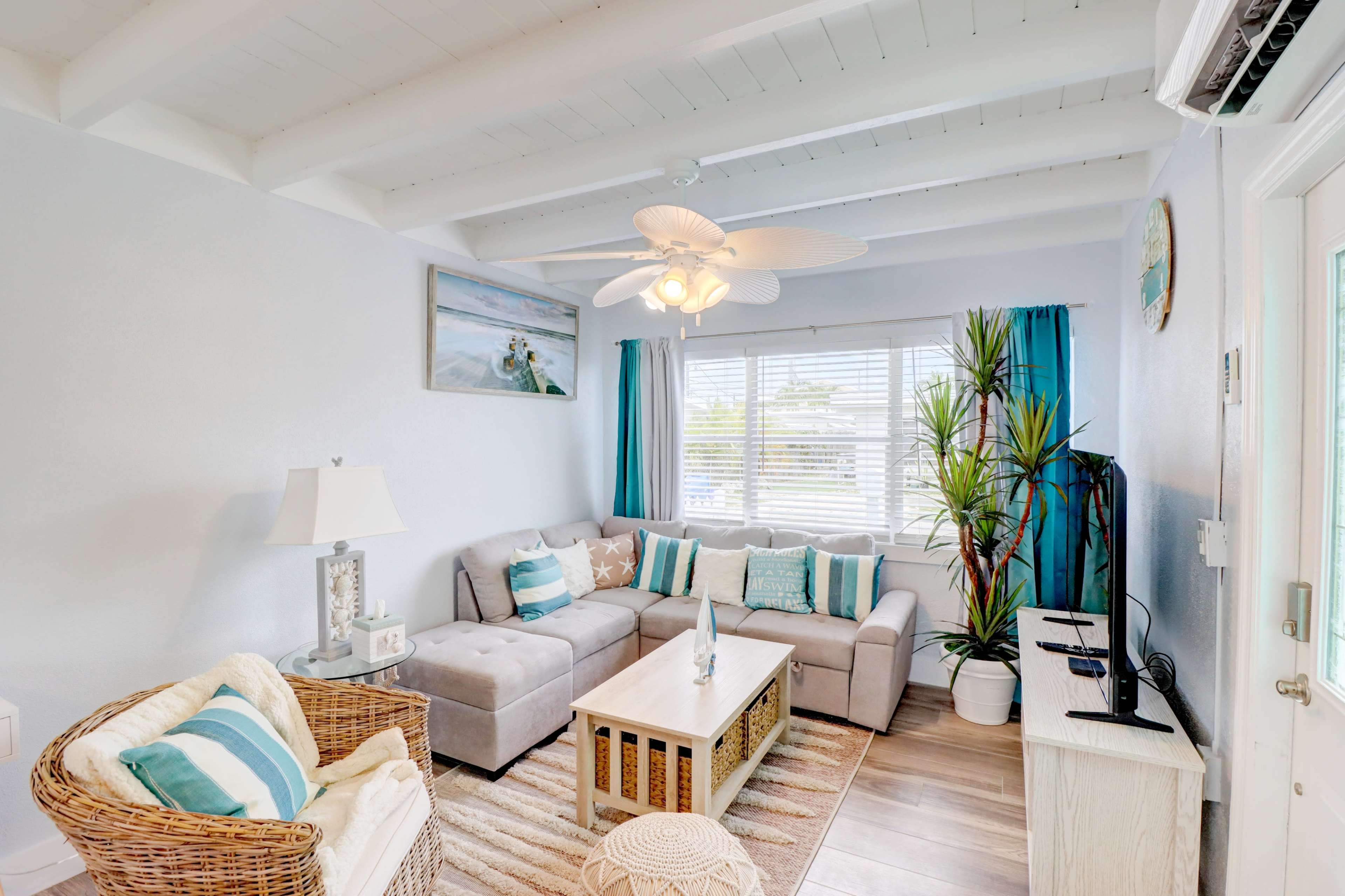 A bright living room with a gray sectional sofa, light wood coffee table, and large window dressed with blue curtains.