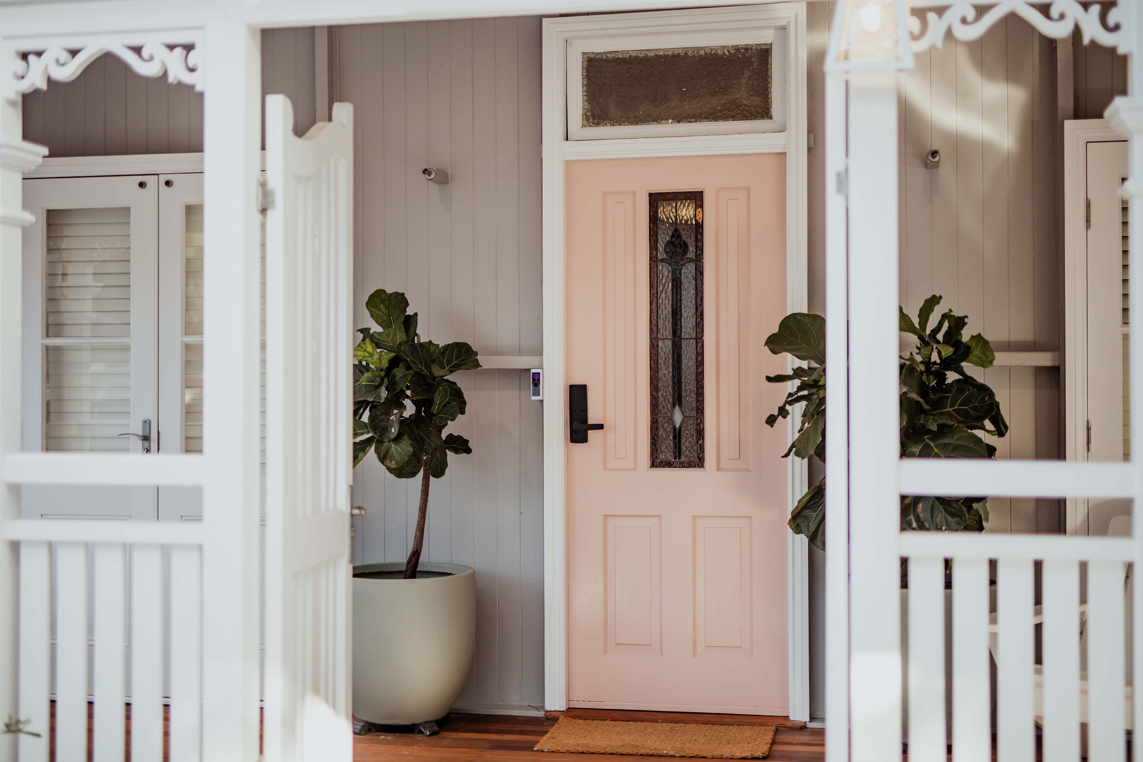 The image shows a front porch with a pink door flanked by potted plants on either side, framed by white, decorative railings.