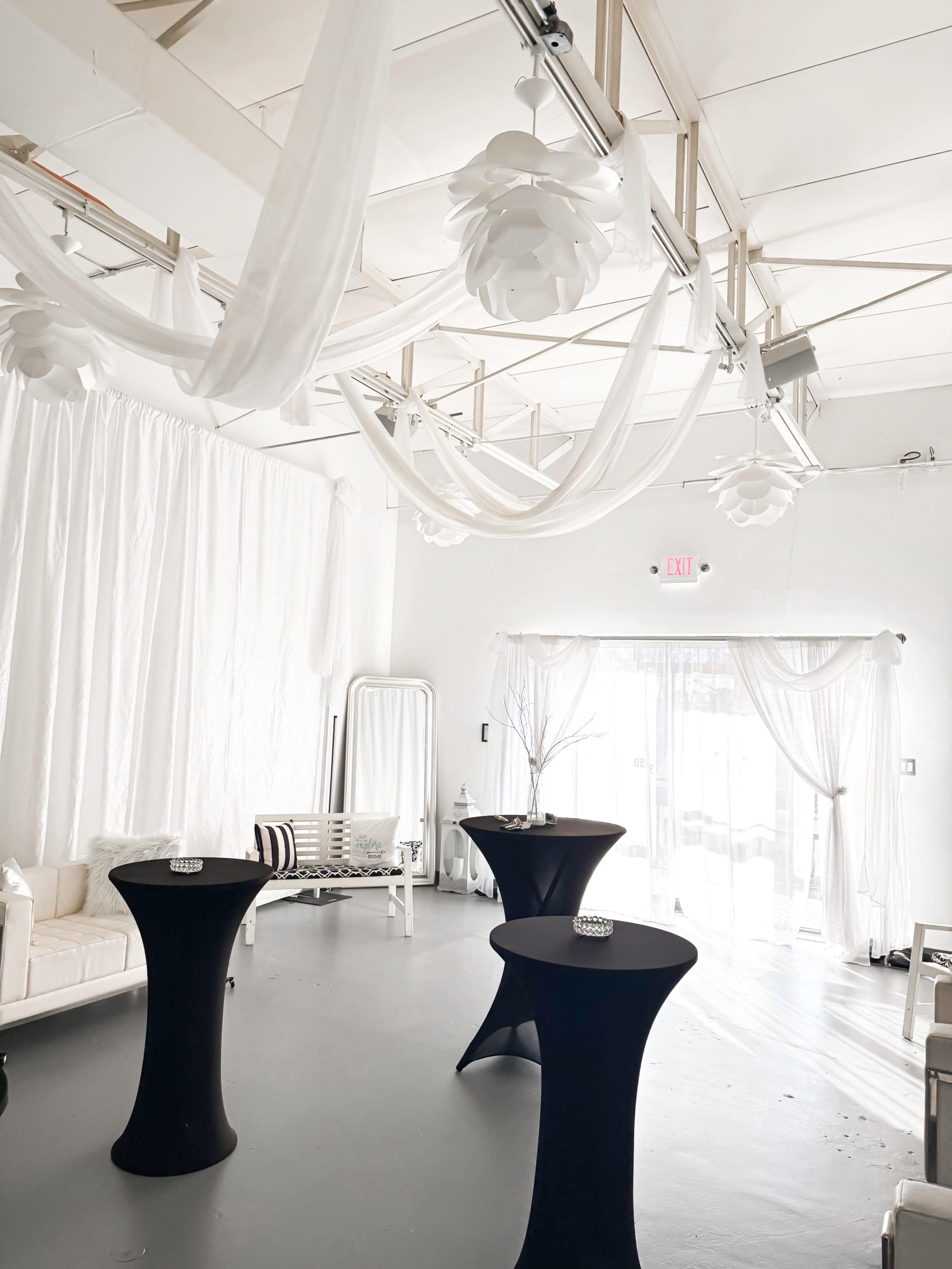 The image shows a bright, minimalist event space with white drapery and two black cocktail tables.