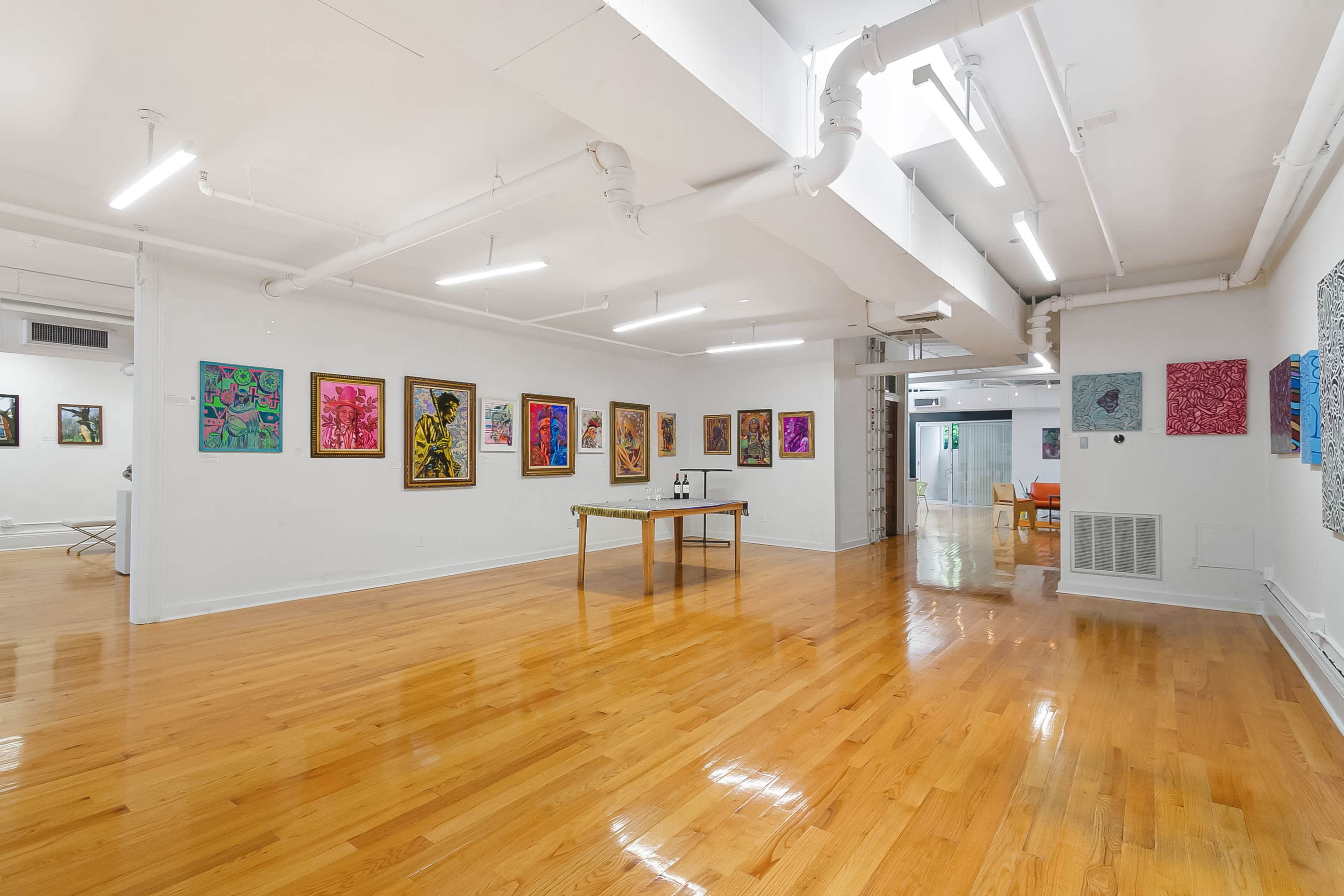 The image shows an art gallery with polished wooden floors, featuring colorful paintings displayed on the walls and a table in the center.