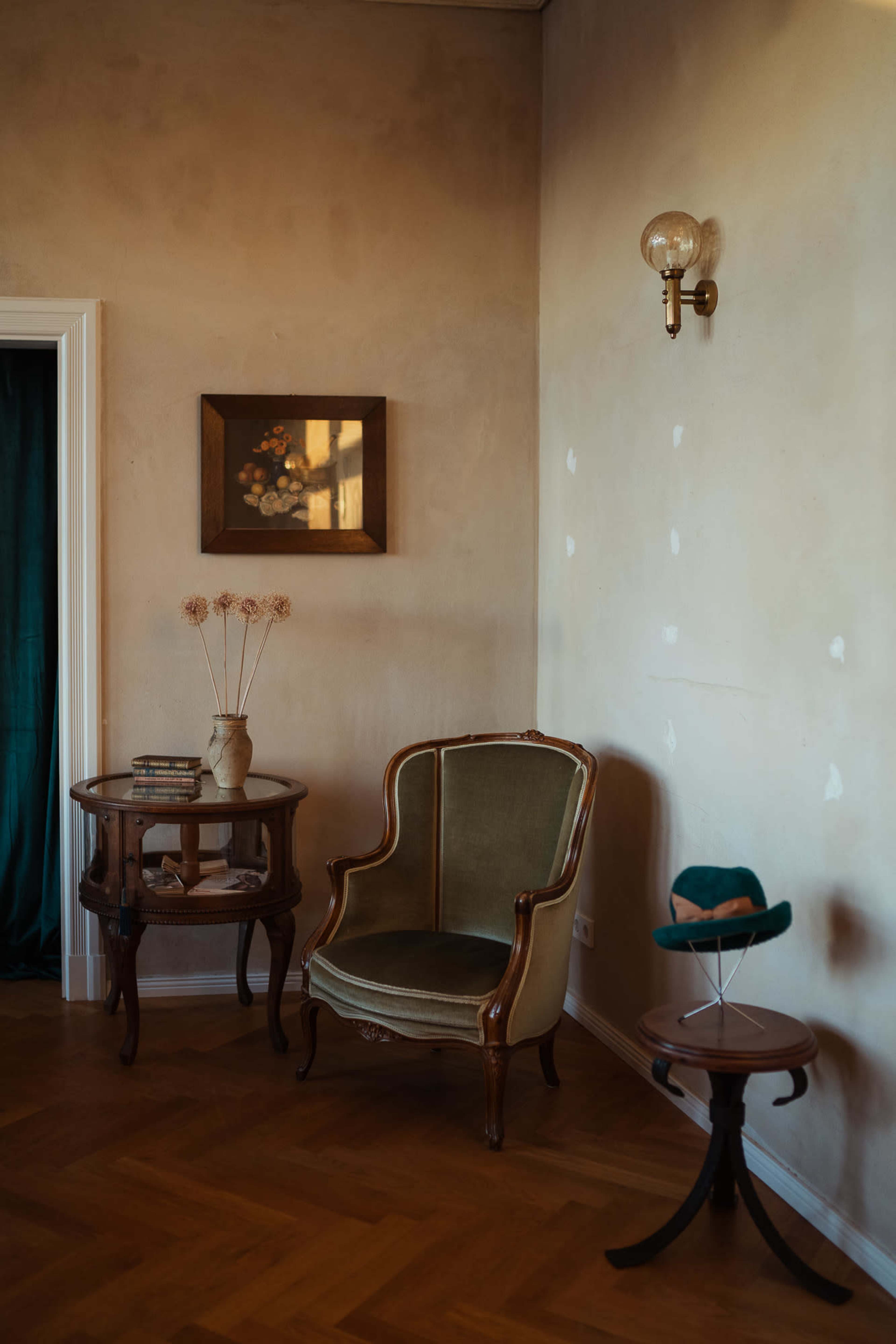 The image shows a corner of a room with a vintage armchair, a small round table with books, a vase of dried flowers, and a piece of framed art on the wall.