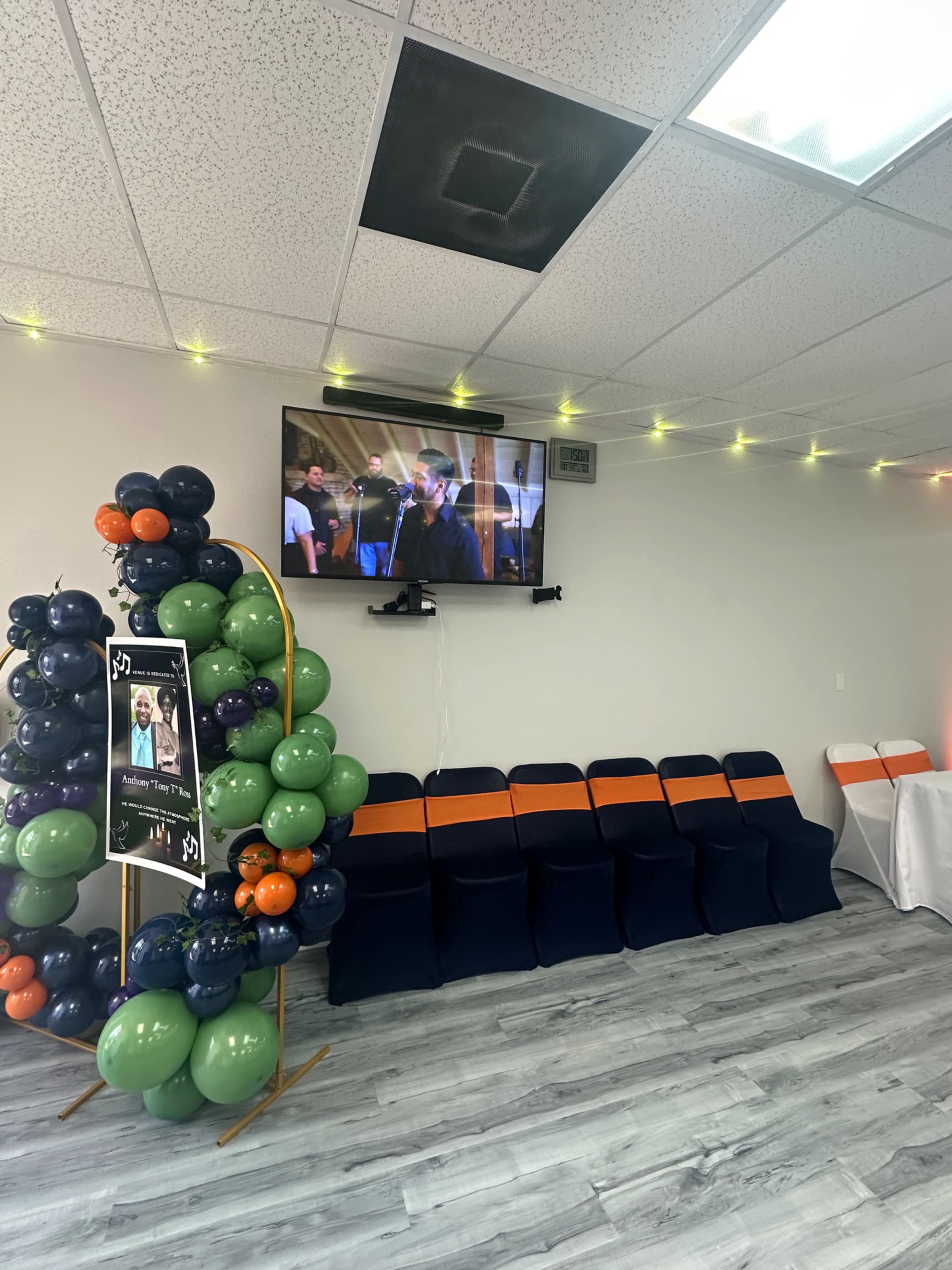 The image shows a decorated room with a green, blue, and orange balloon arch, a television displaying a performance, and rows of chairs.