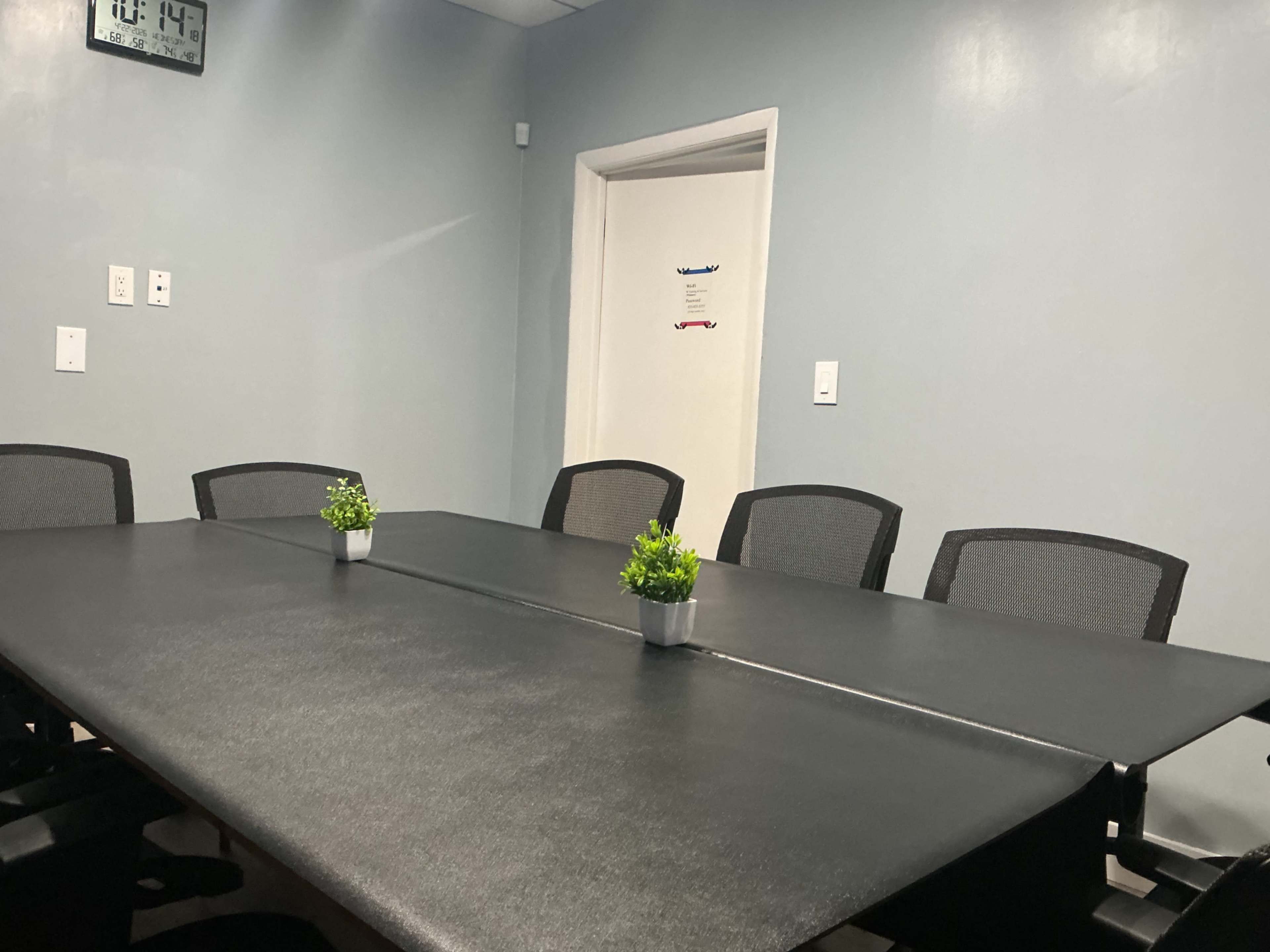 The image shows a conference room with a large black table surrounded by mesh chairs and small potted plants on the table.