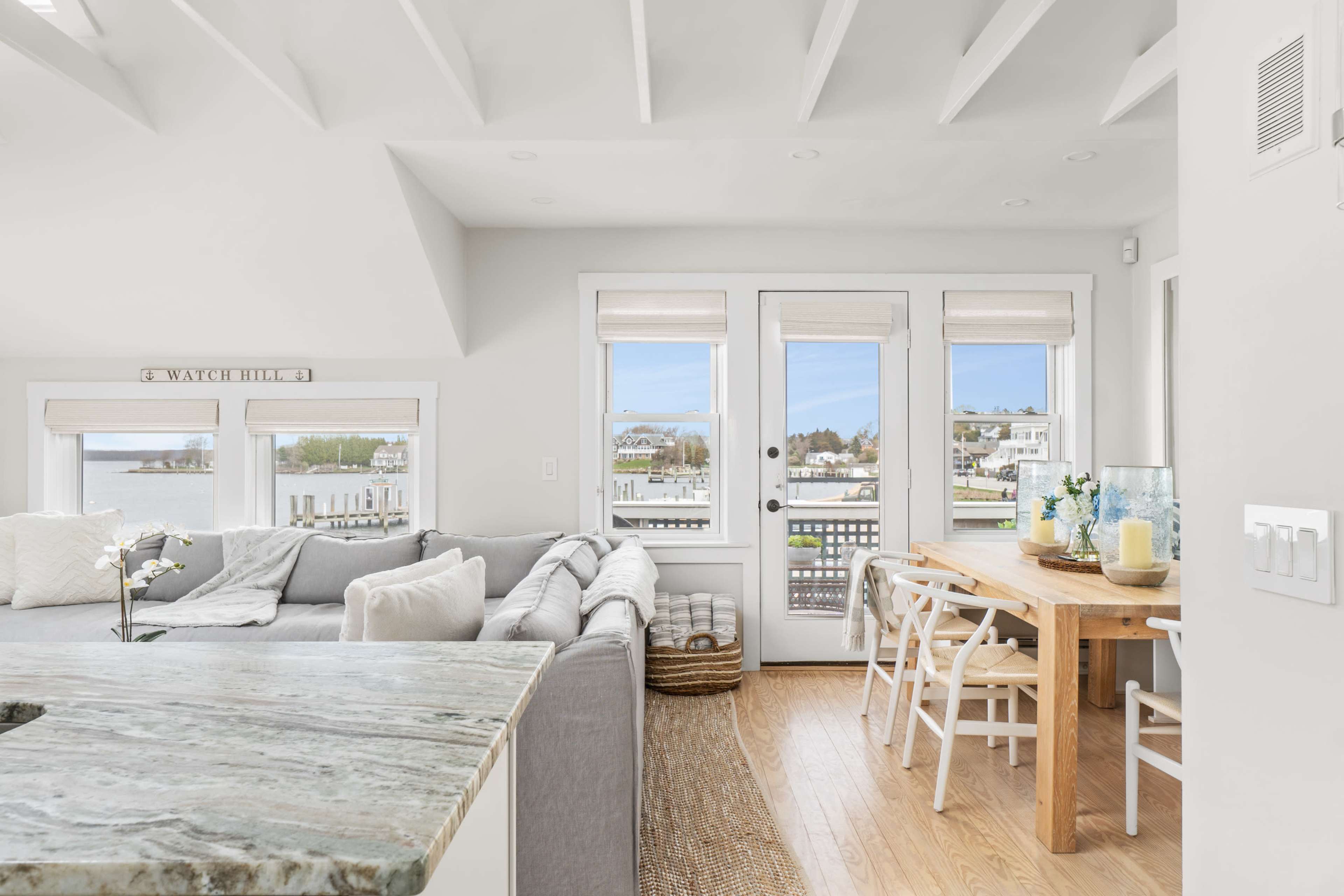 The image shows a light-filled living space with a grey sofa, a wooden dining table, and large windows overlooking a water view.